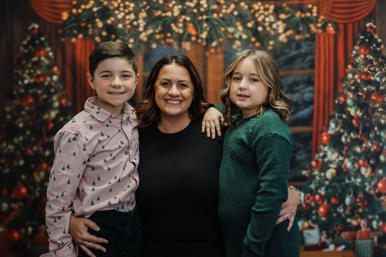 A woman smiles with two children in front of decorated Christmas trees in a festive holiday portrait.
