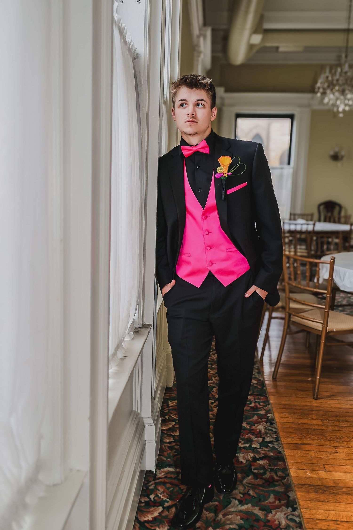 Young man in black tuxedo with hot pink vest and bow tie stands by window in elegant event venue.