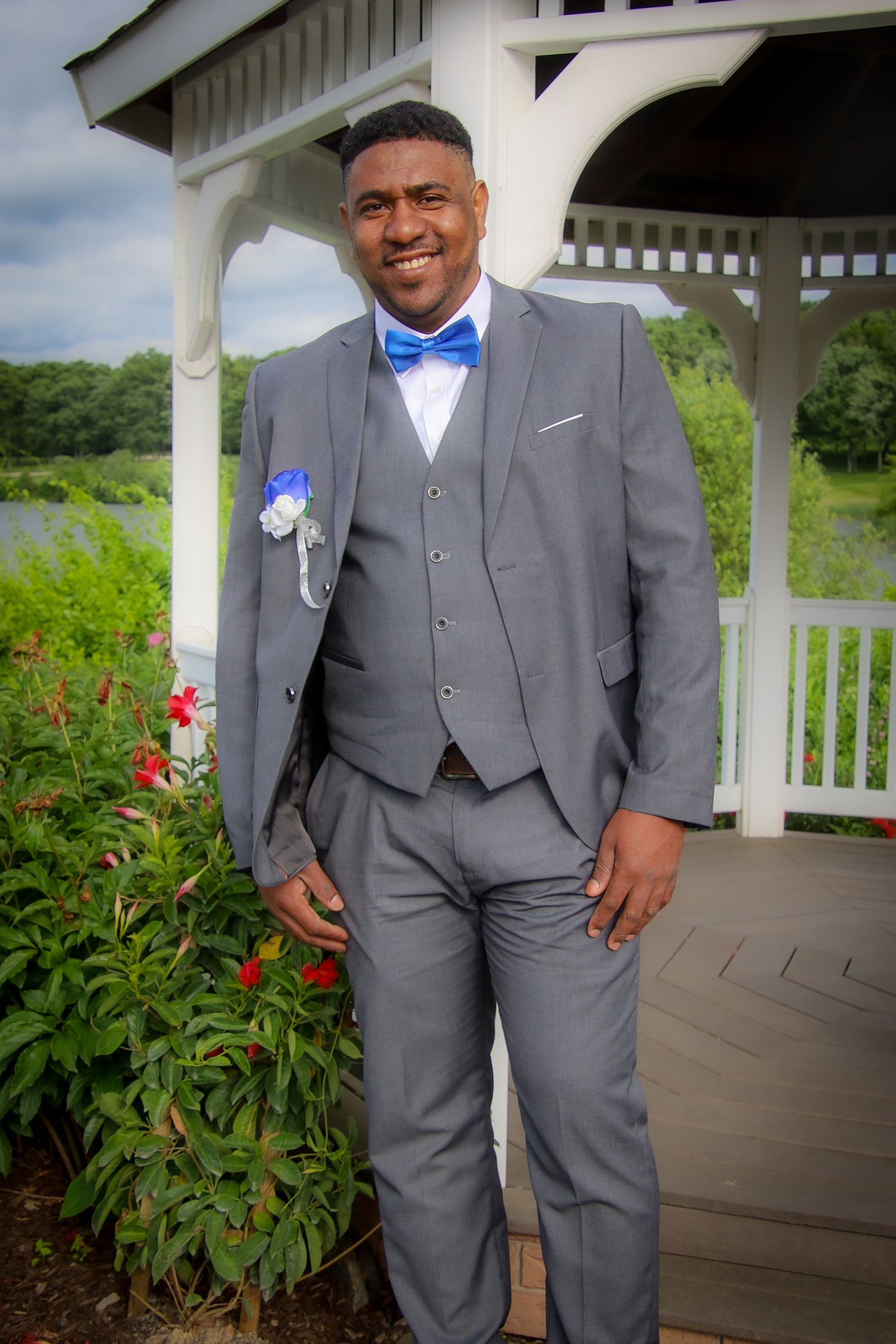 A person in a gray three-piece suit and blue bow tie poses in front of a white gazebo surrounded by flowers.