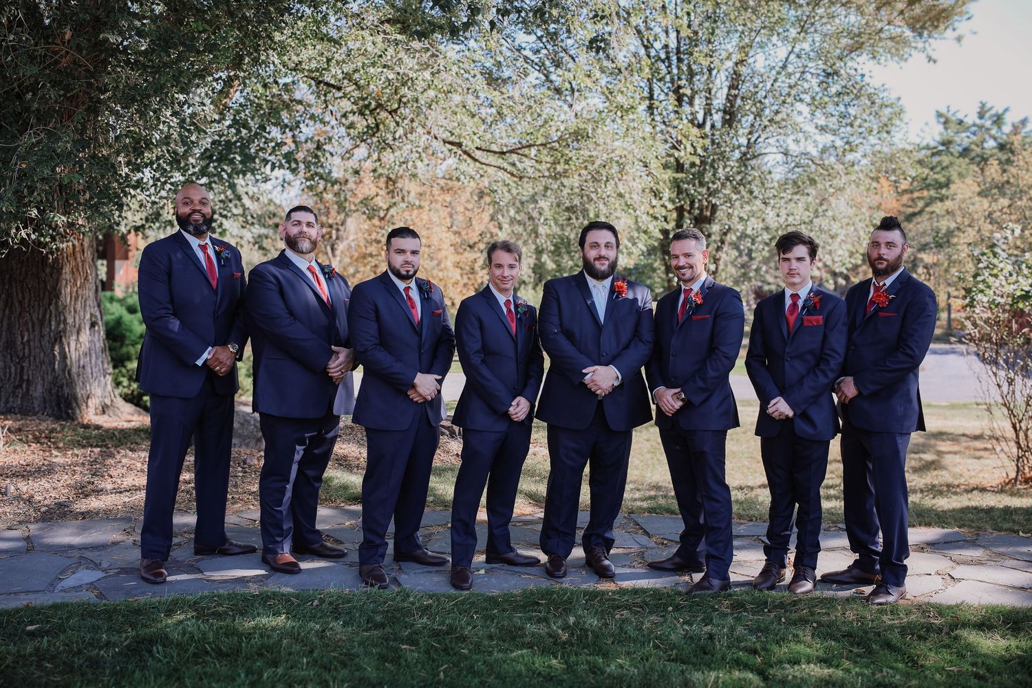 Wedding party wearing navy suits and red ties pose together in a garden setting for formal group photos.