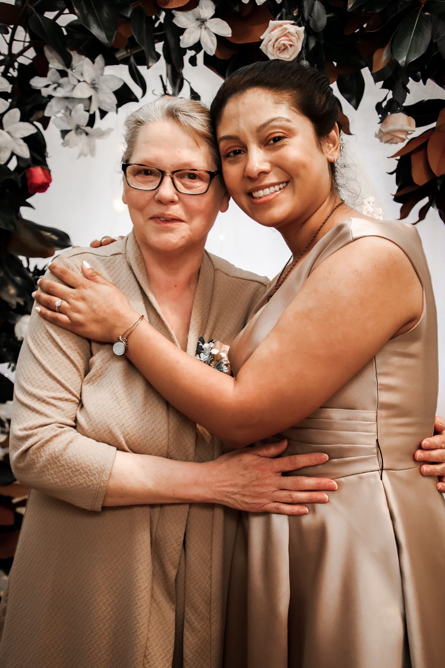 Two people in beige dresses sharing a warm embrace against a floral backdrop.