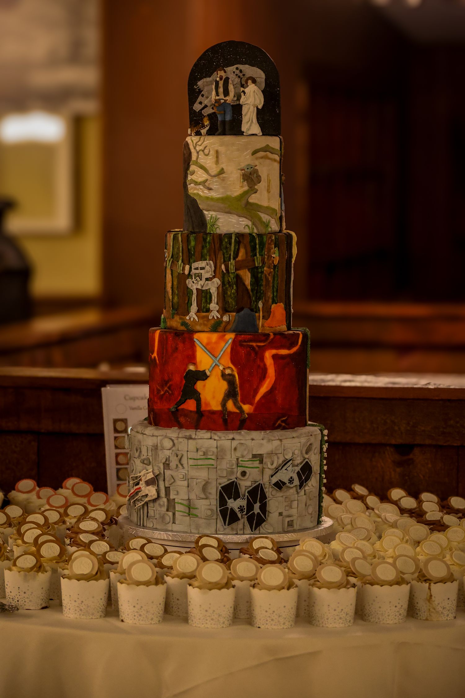 A tiered artistic wedding cake with painted designs and cupcakes arranged around the base on a white tablecloth.