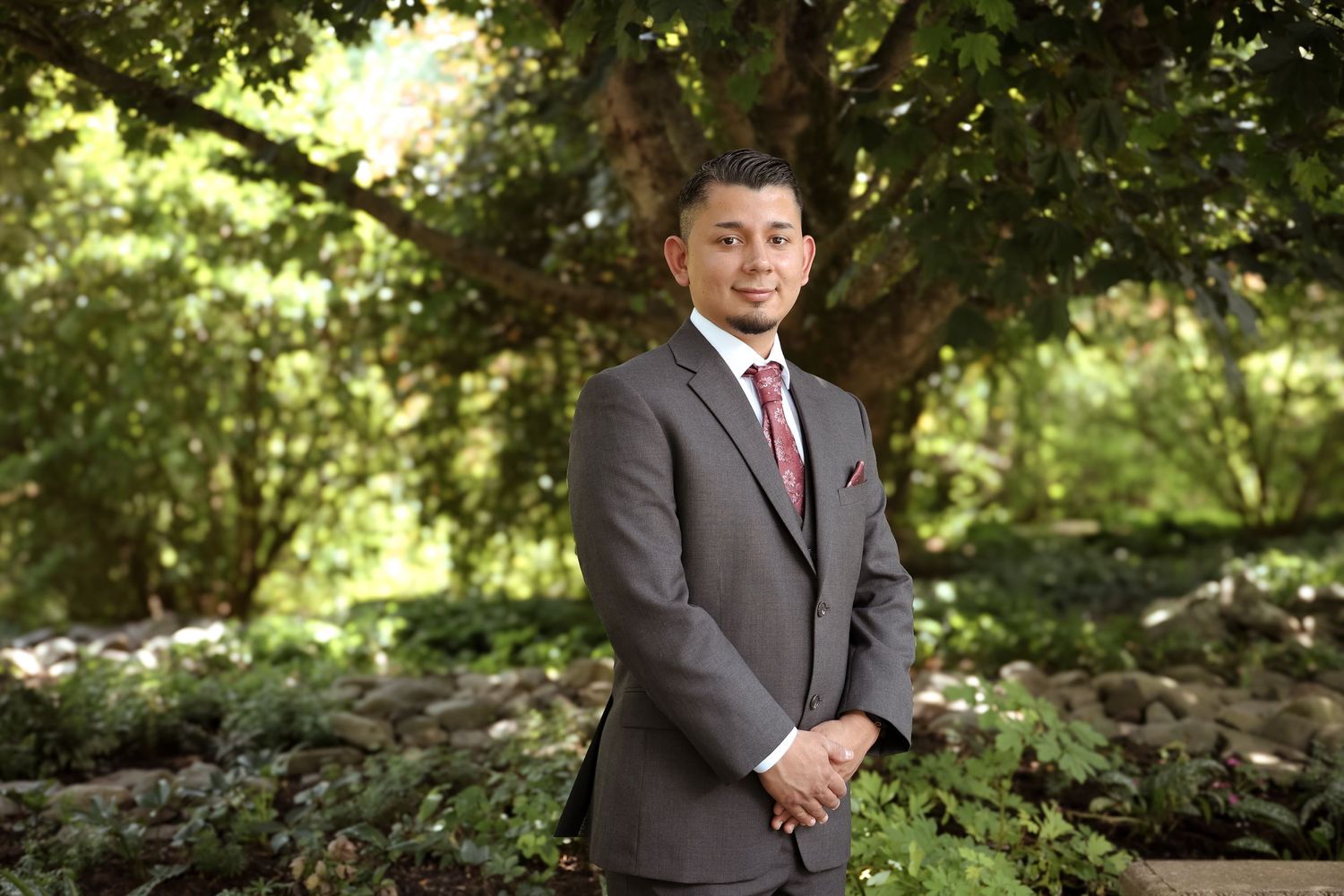 Professional portrait of someone in a gray suit and red tie standing outdoors with trees and natural scenery in background.
