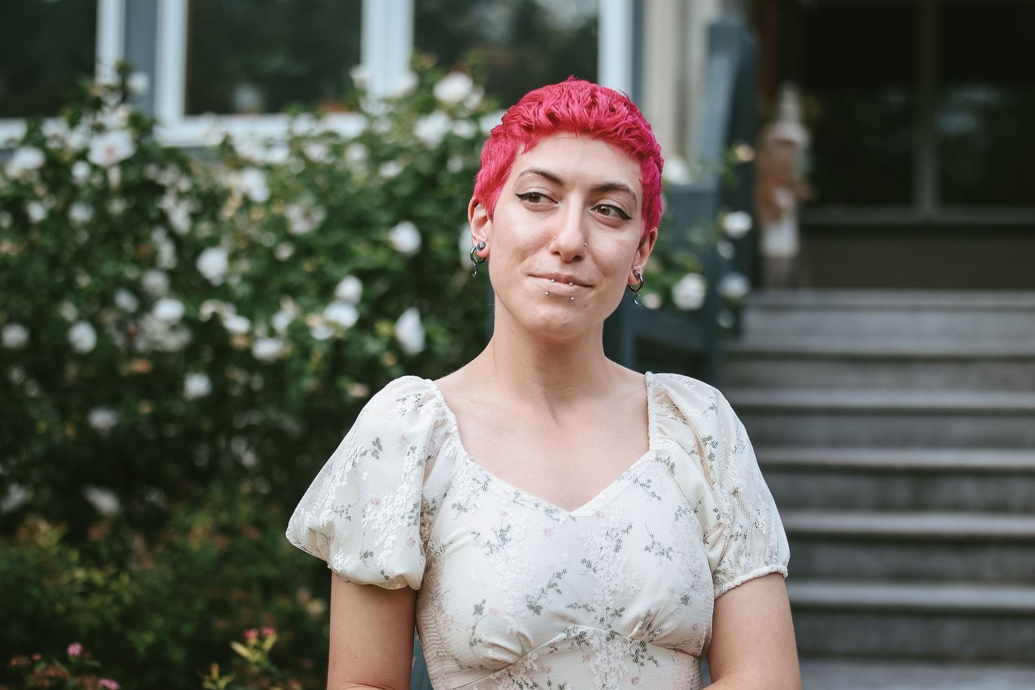 A person with bright pink hair wearing a white floral dress poses in front of a garden setting.