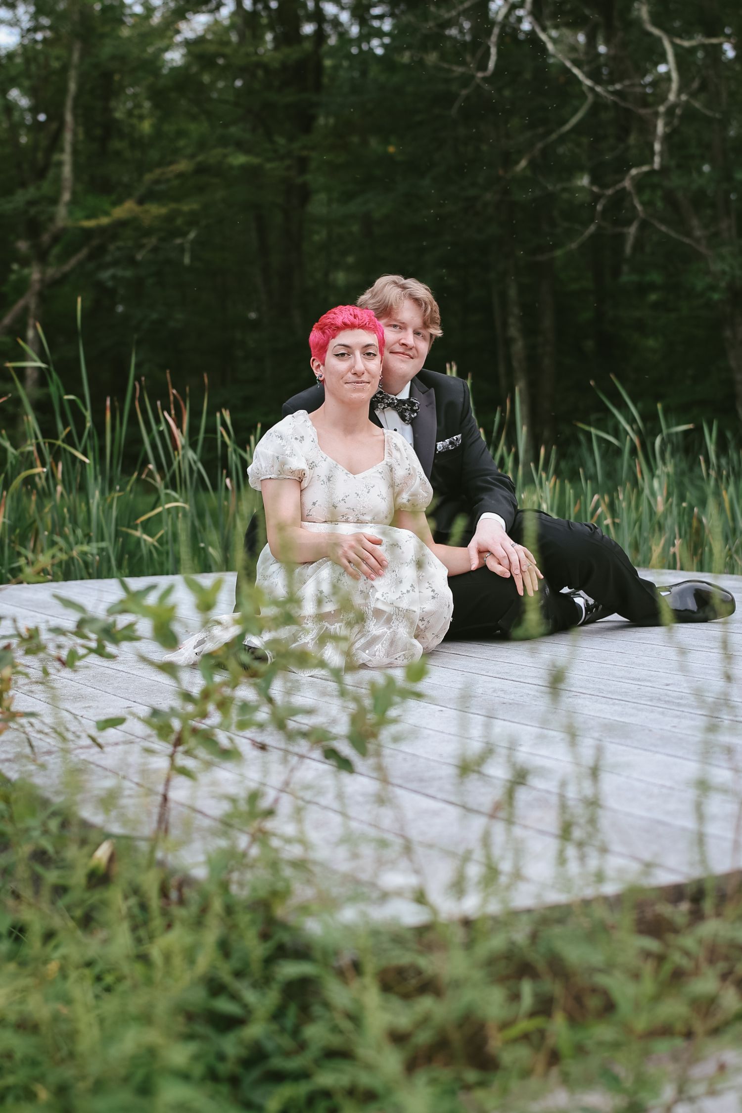 A couple in formal attire sits together on a wooden boardwalk surrounded by lush greenery and tall grass.