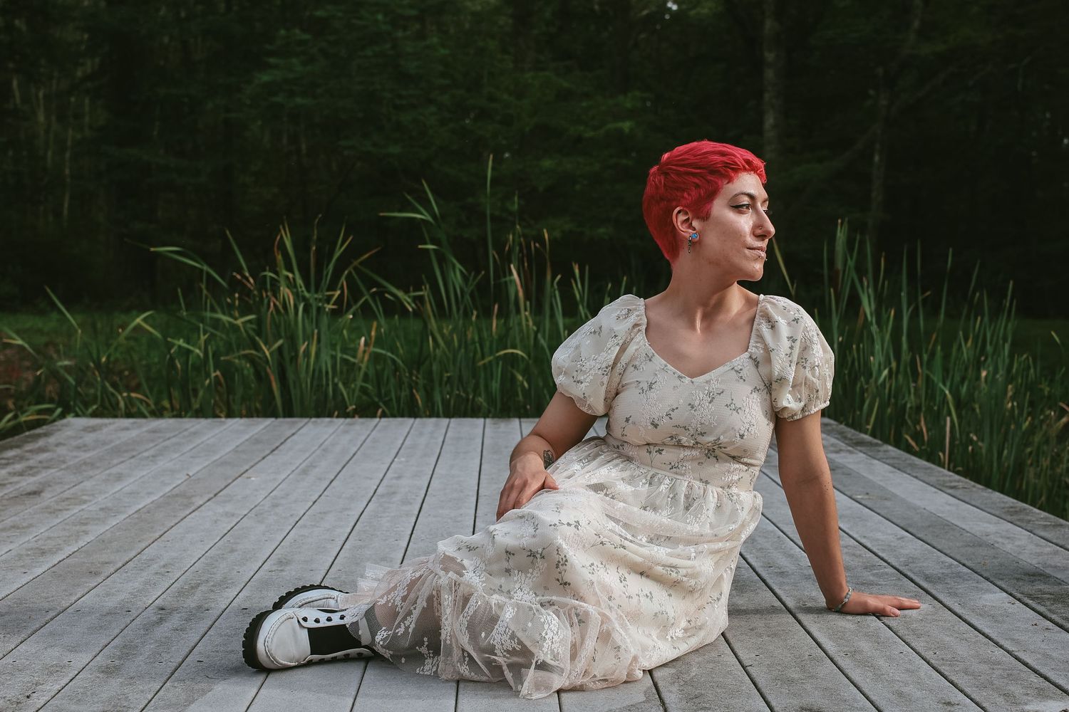 A person in a lace dress lies on a wooden boardwalk surrounded by tall grass and trees.