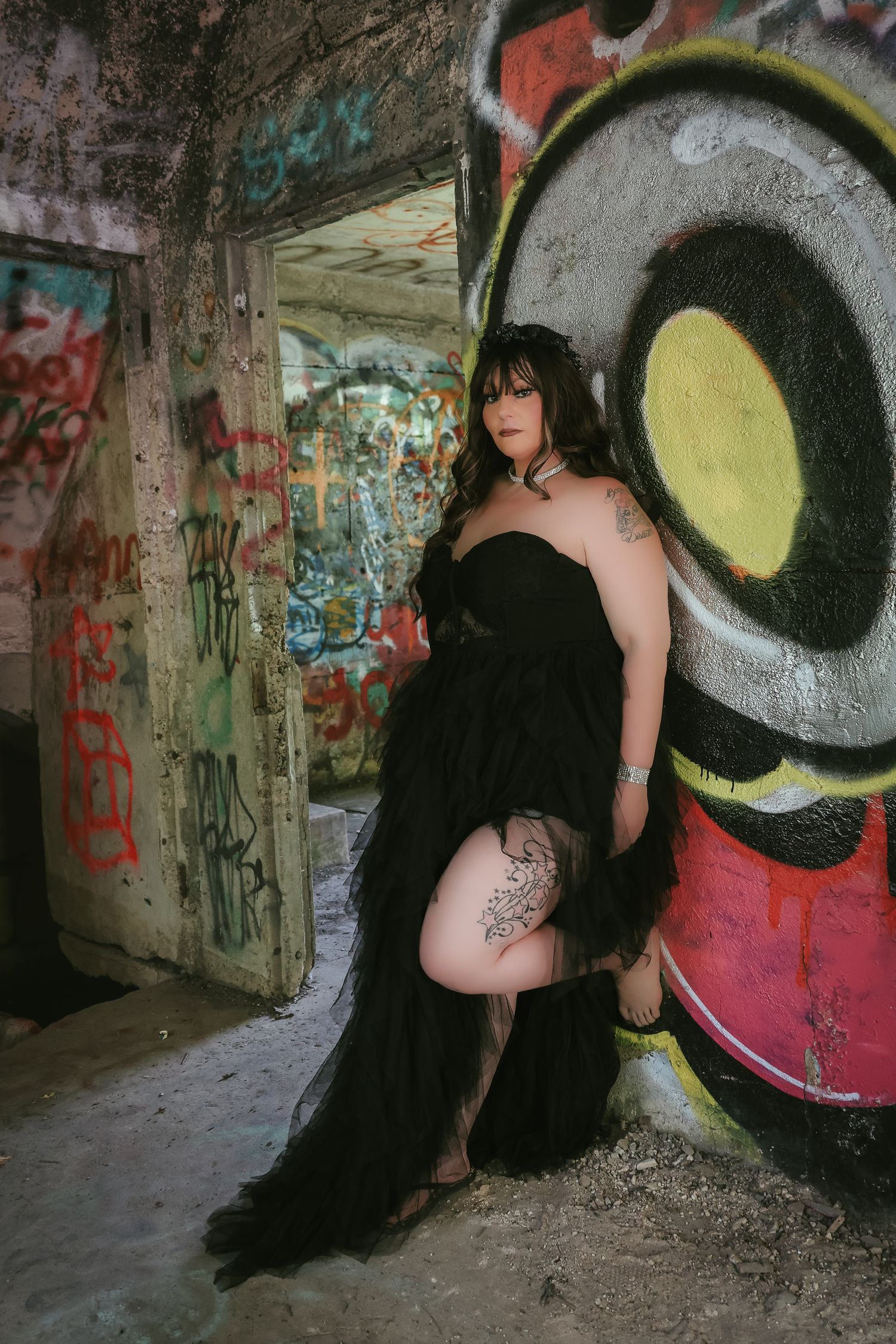 A figure in a flowing black dress poses against graffiti walls in an urban tunnel setting.