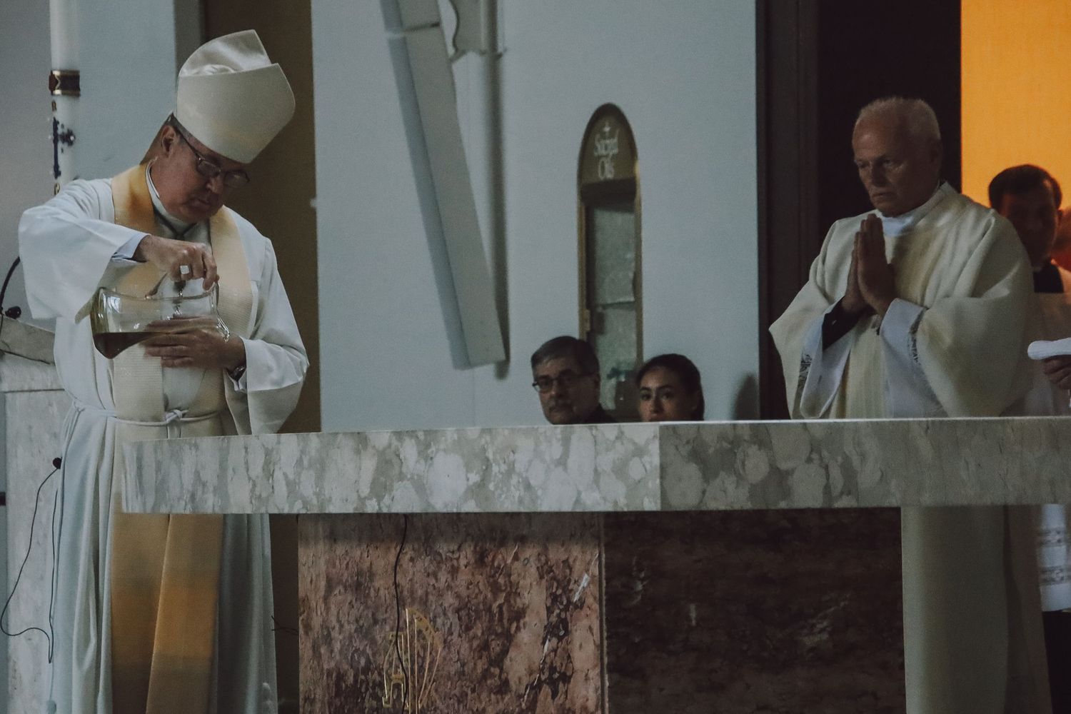 Catholic priests in white robes gather at the altar during a religious ceremony with an orange wall backdrop.