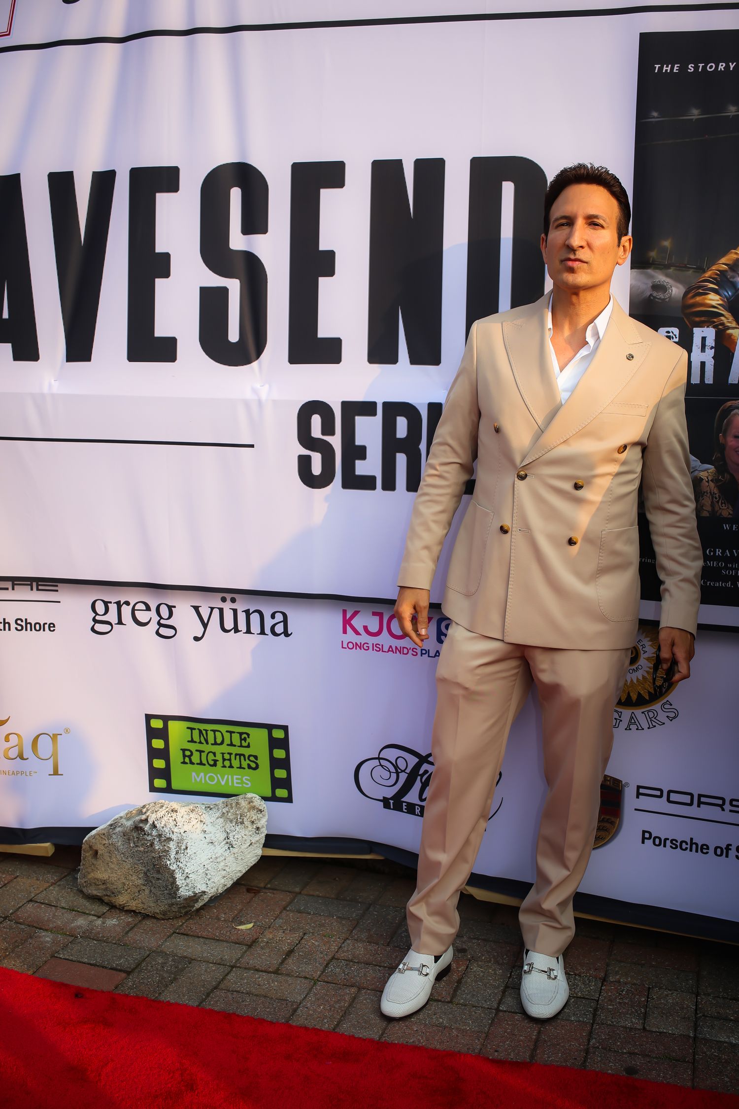 A person poses on a red carpet wearing a beige double-breasted suit with white sneakers at a premiere event.