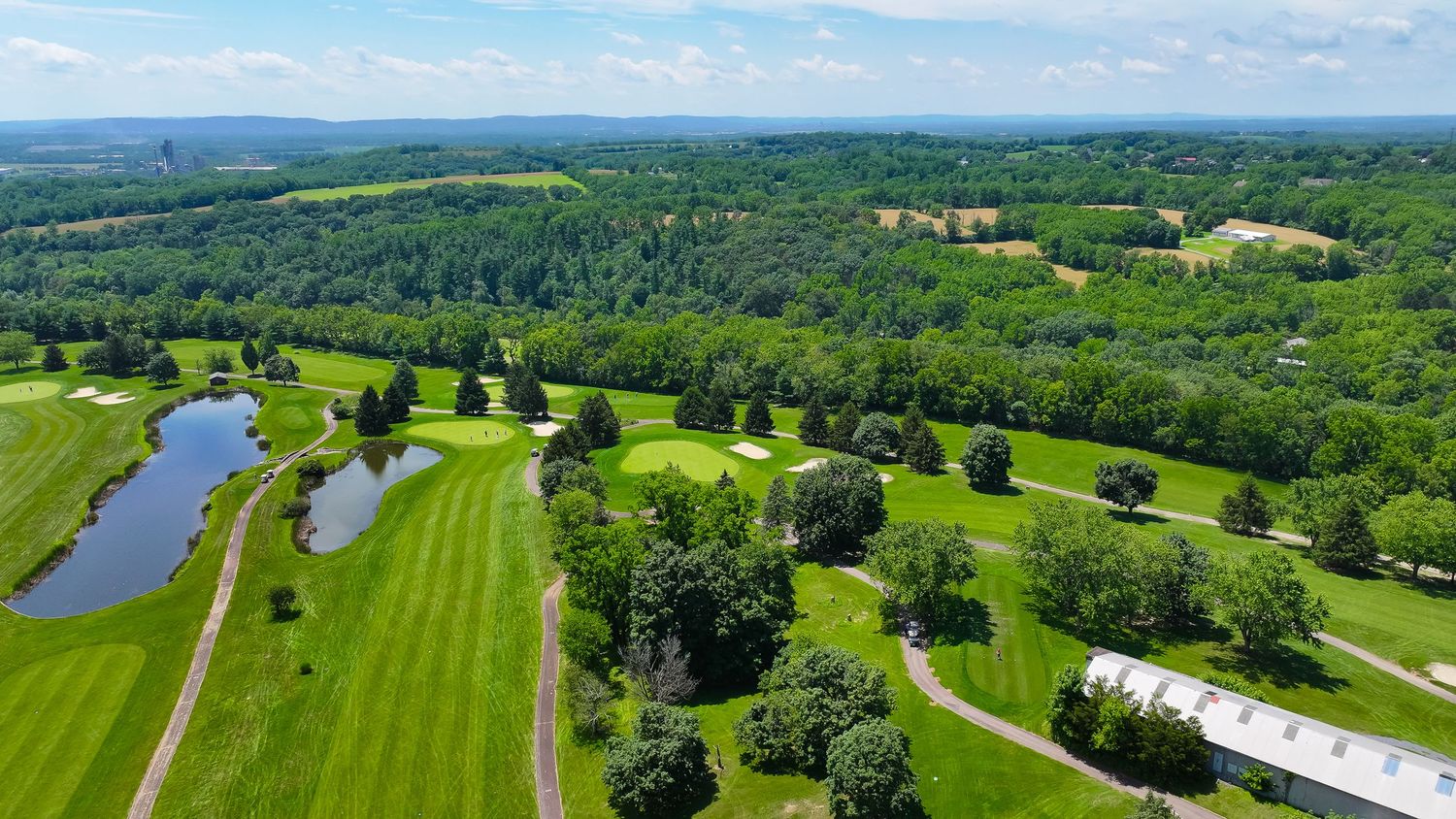 Aerial panoramic view of a lush golf course with rolling greens, ponds, and tree-lined fairways on a sunny day.