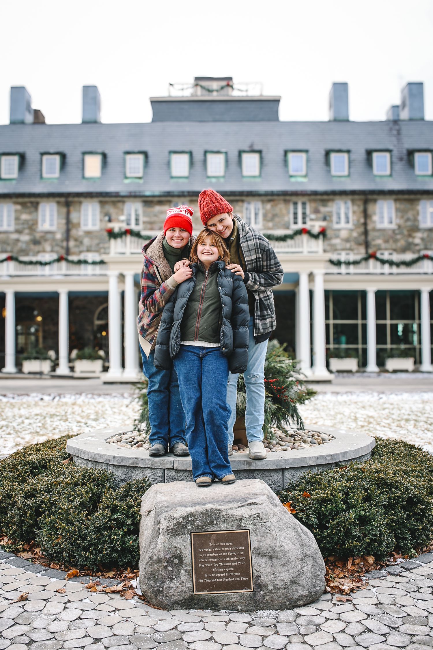 Family in winter clothing poses together on a commemorative stone marker outside a historic building with colonial architecture.