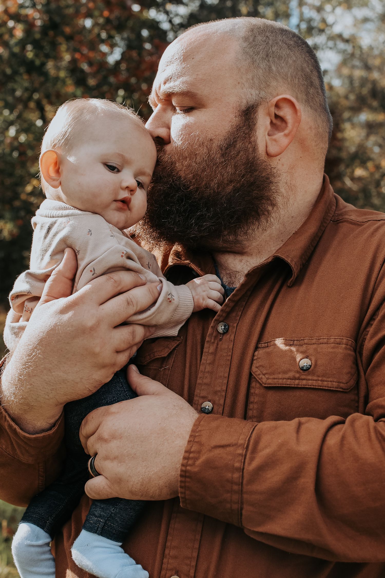 A tender moment between a bearded adult in a brown jacket holding and kissing a small baby in light colored clothing.