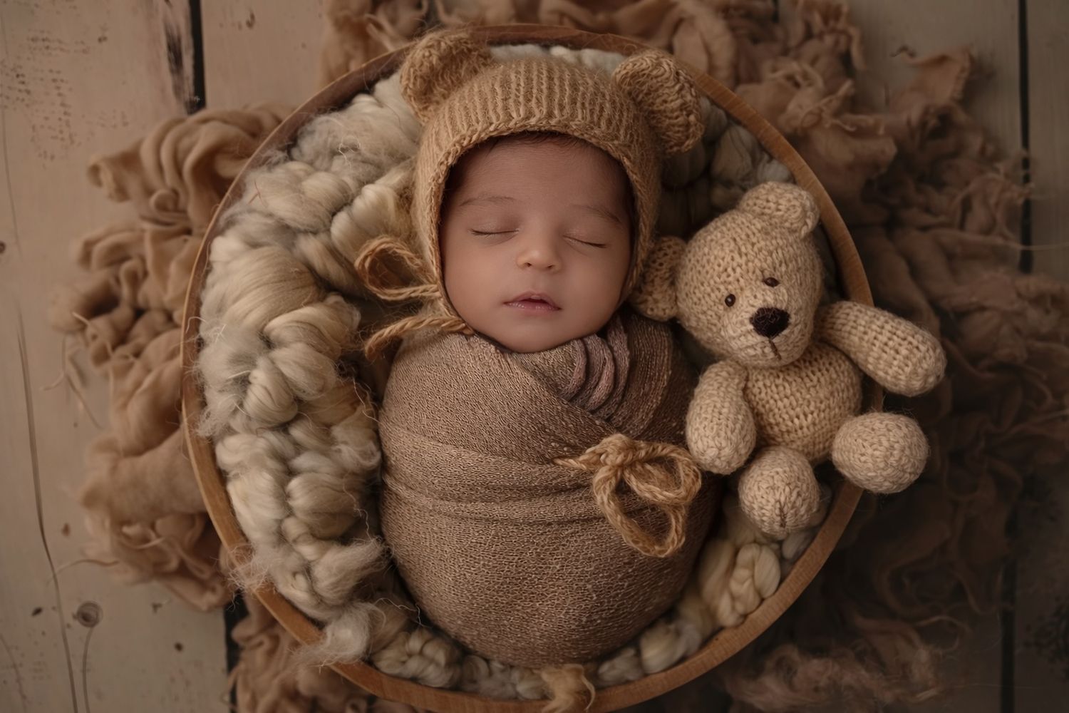 Sleeping baby wrapped in brown knit swaddle with matching bonnet lies in basket next to plush teddy bear.