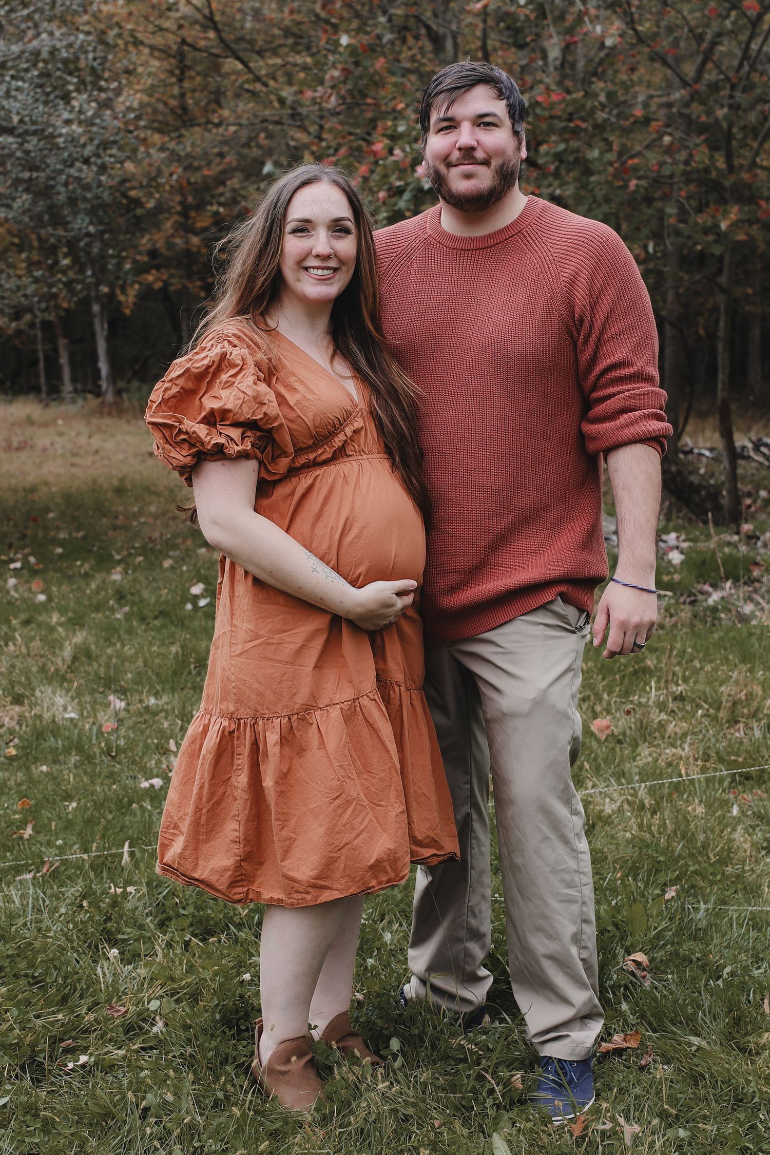 A couple in autumn attire stands outdoors, with the woman wearing a rust-colored dress and cradling her pregnant belly.