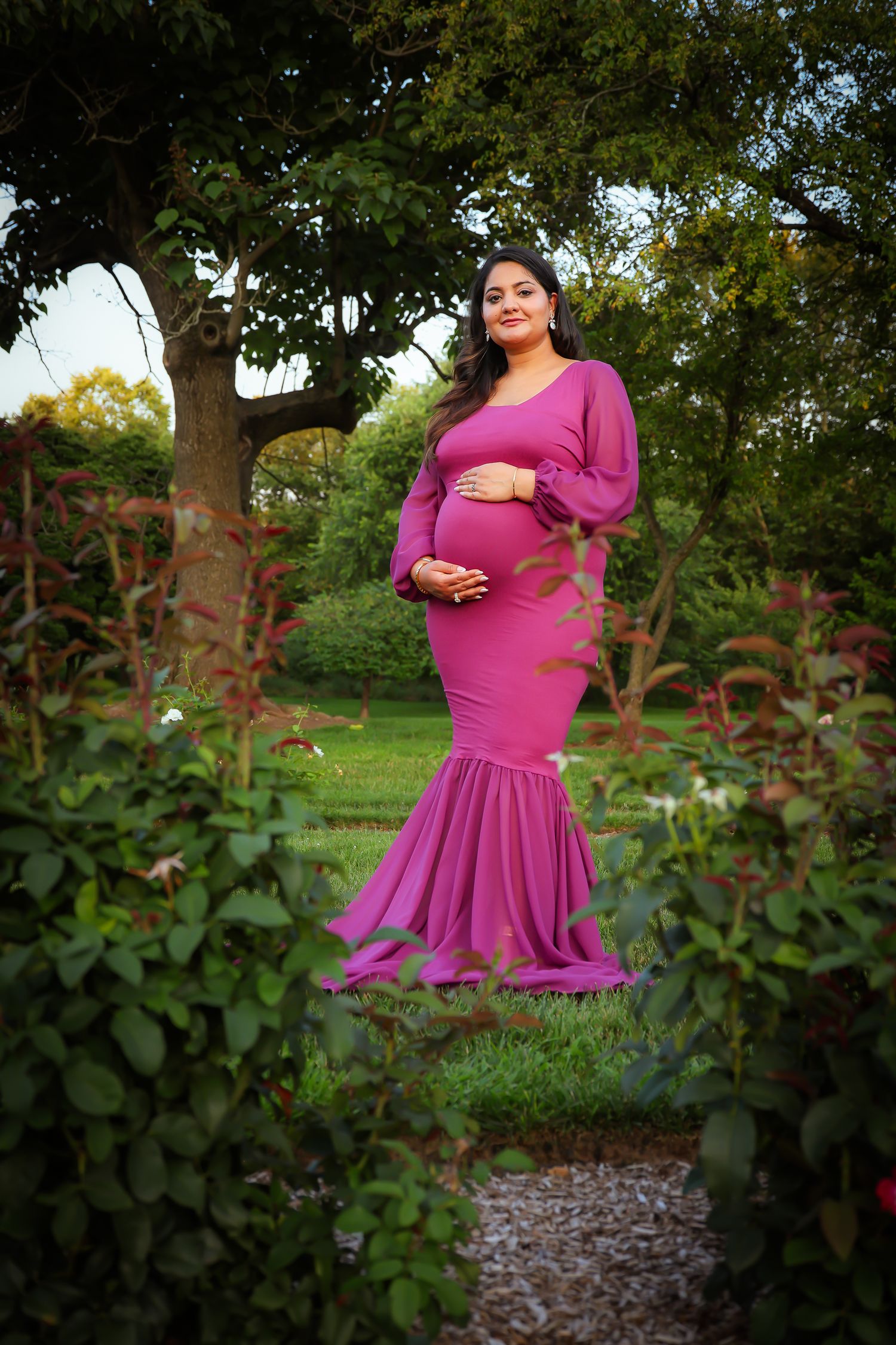 A bright fuchsia dress flows elegantly in an outdoor garden setting with lush greenery and trees in the background.