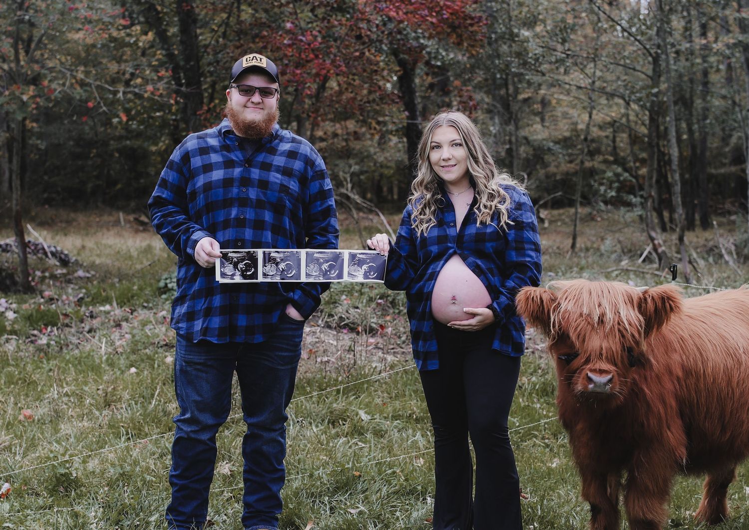 A couple stands together in a field with a Highland cow during a rustic outdoor photo session.