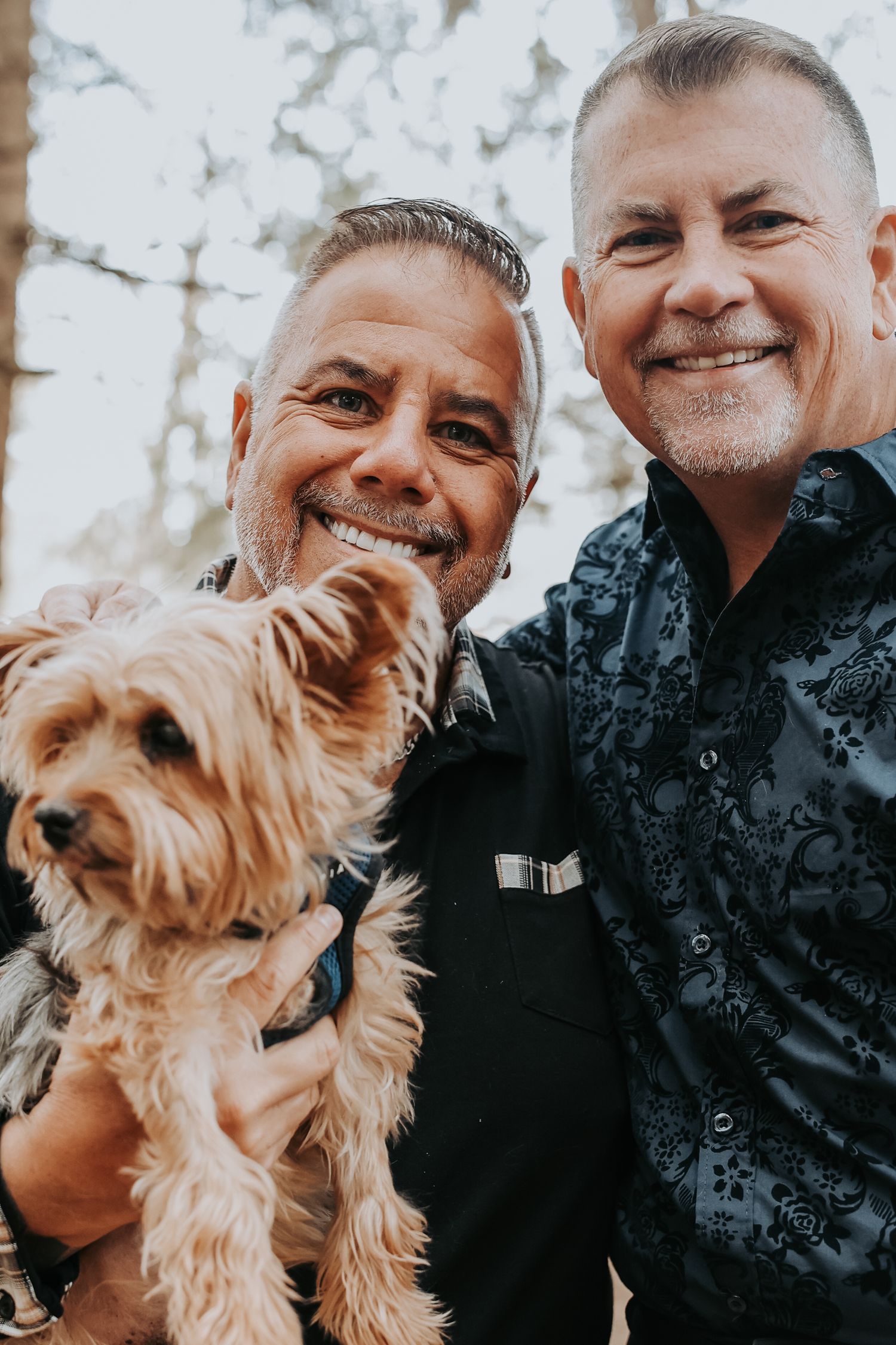 A happy couple poses outdoors with their small Yorkshire Terrier dog on a bright day.