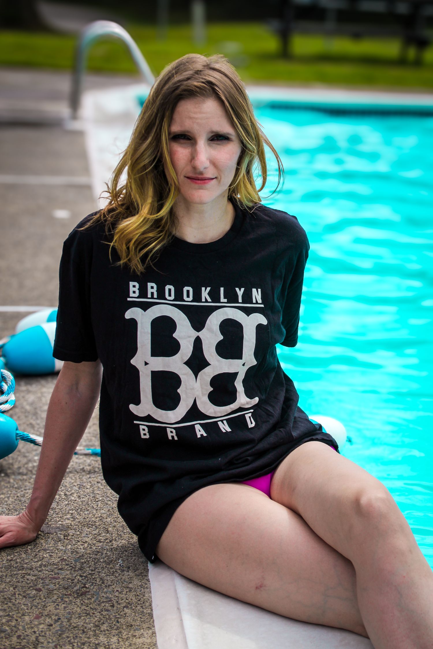 Person lounging by bright blue swimming pool wearing black Brooklyn t-shirt on sunny summer day.