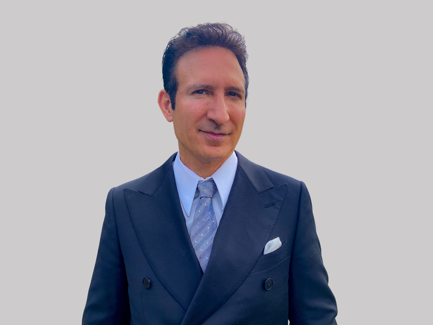 A person in a dark business suit and light purple tie poses confidently against a gray background.