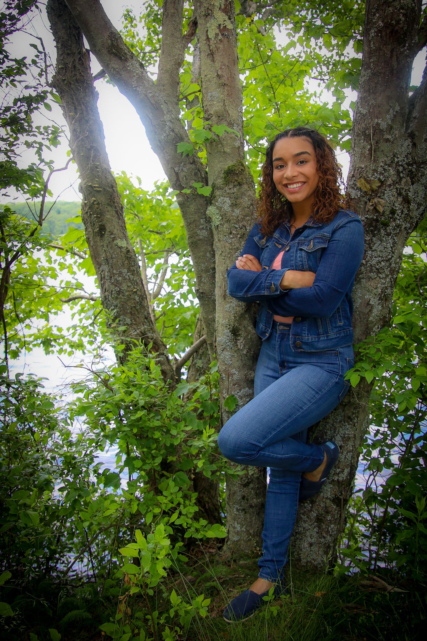 A person wearing a denim jacket and jeans sits casually perched in a tree surrounded by green foliage.