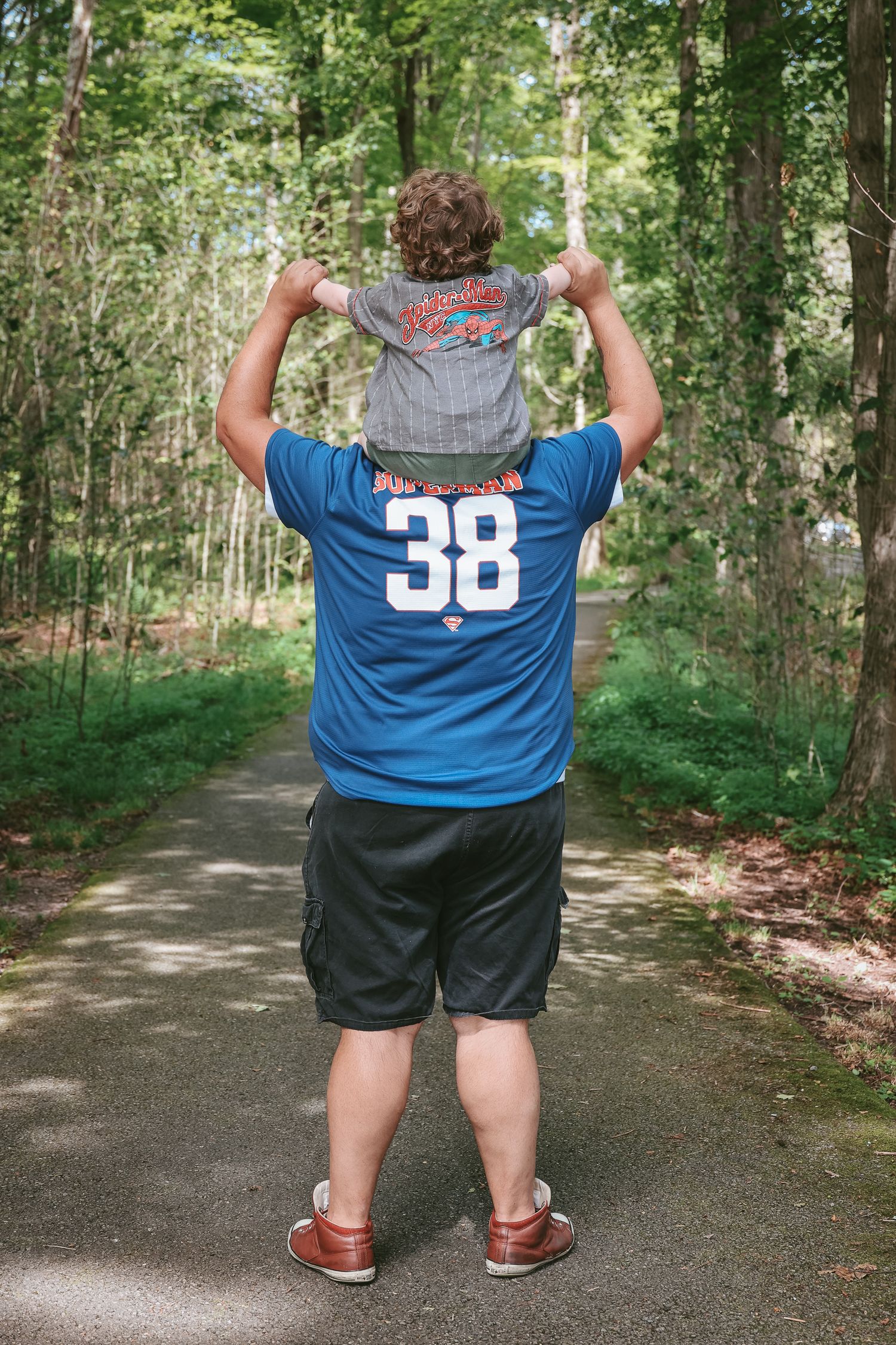 Someone wearing a blue jersey with number 38 carries a small child on their shoulders while walking on a forest path.