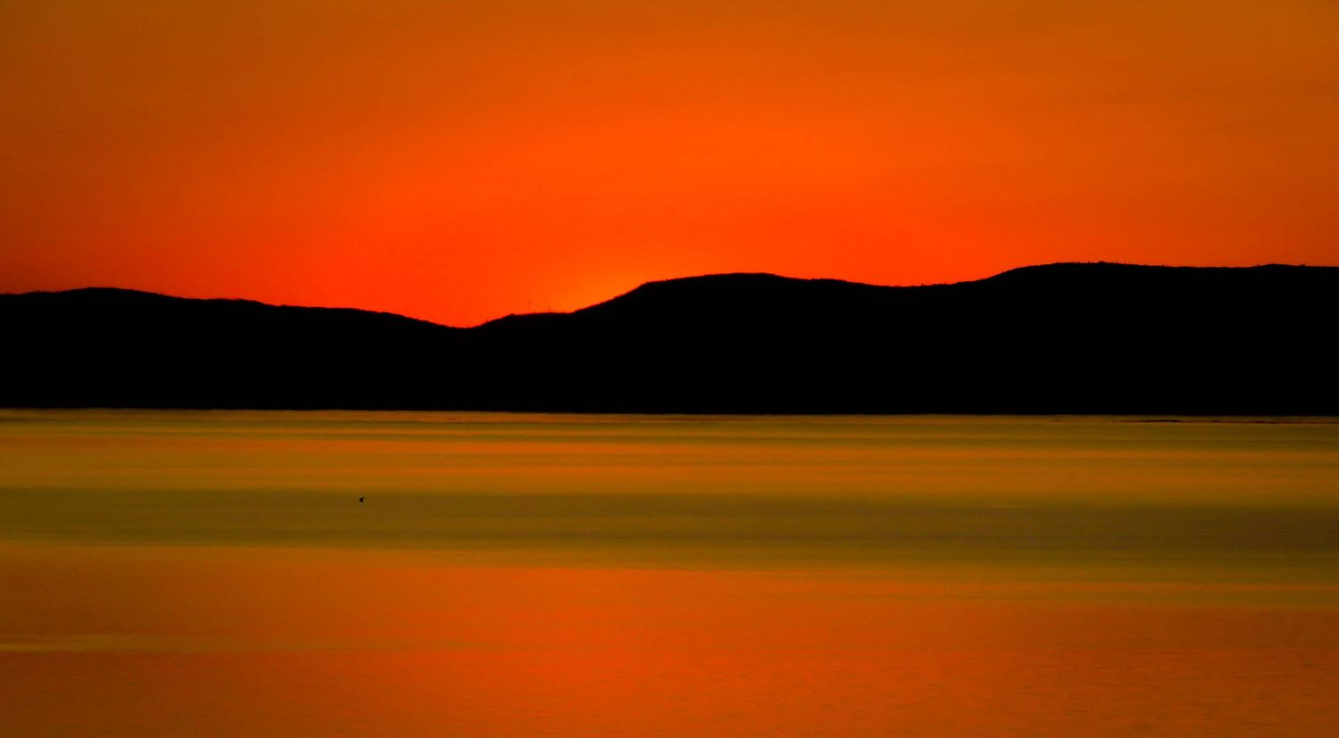 Minimalist landscape of orange sunset behind dark mountain ridges reflected on still water.