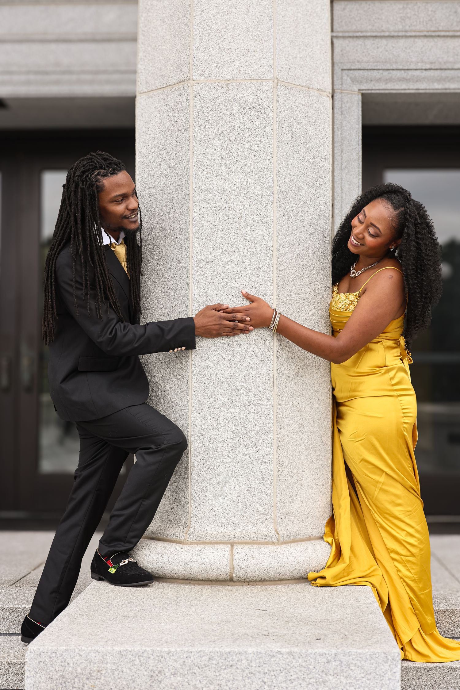A couple in contrasting black and yellow outfits pose playfully around a white stone column in a photo series.