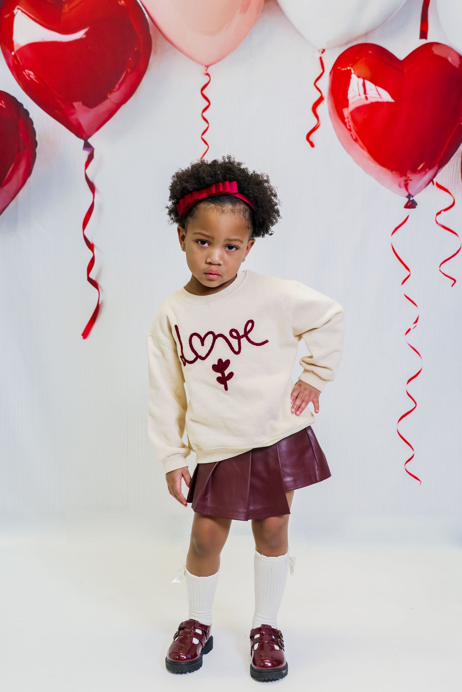 A toddler poses in a cream sweater and burgundy shoes against a romantic backdrop of heart-shaped balloons.
