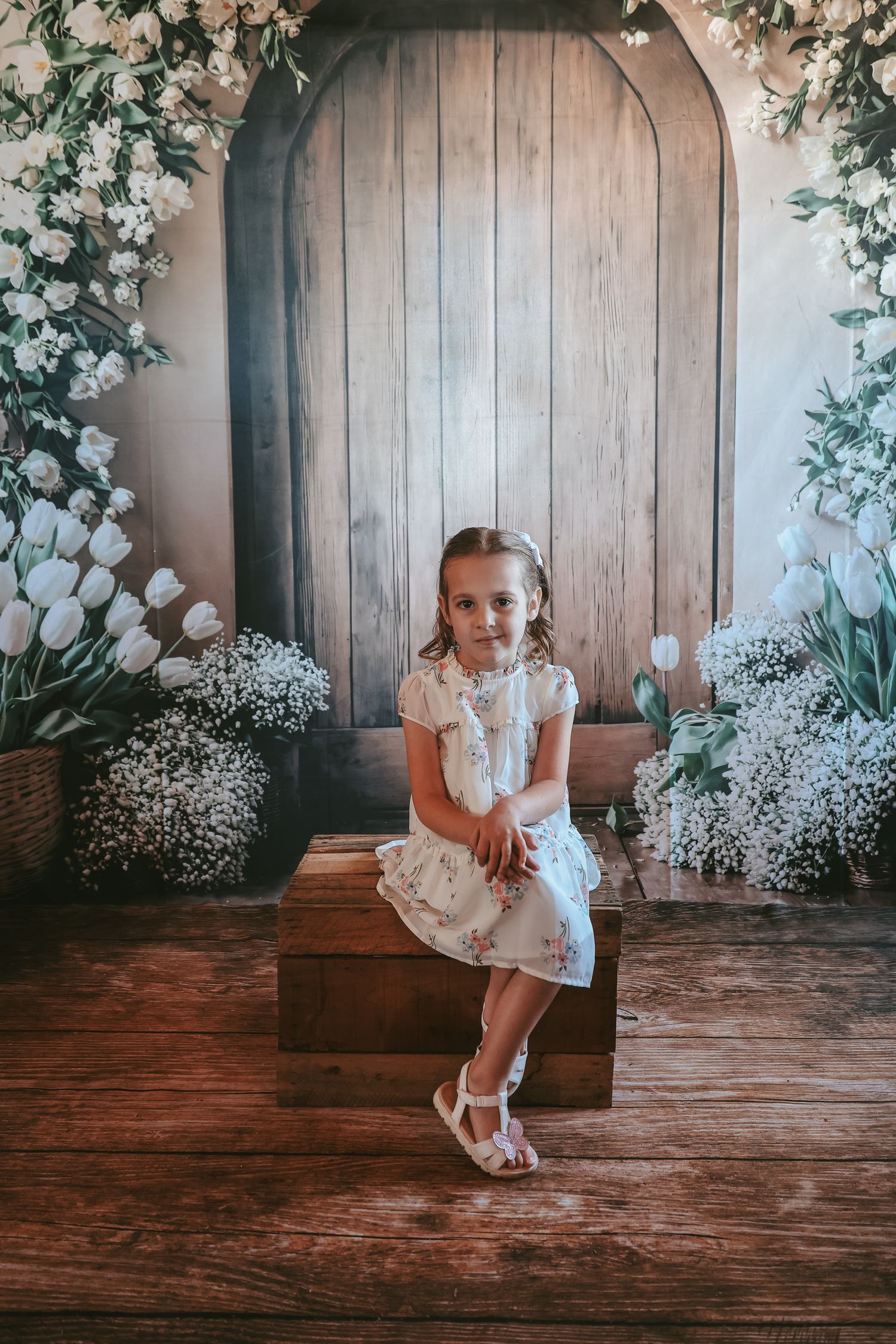 A child sits on a wooden box against a rustic door backdrop with white floral decorations and baby's breath arrangements.