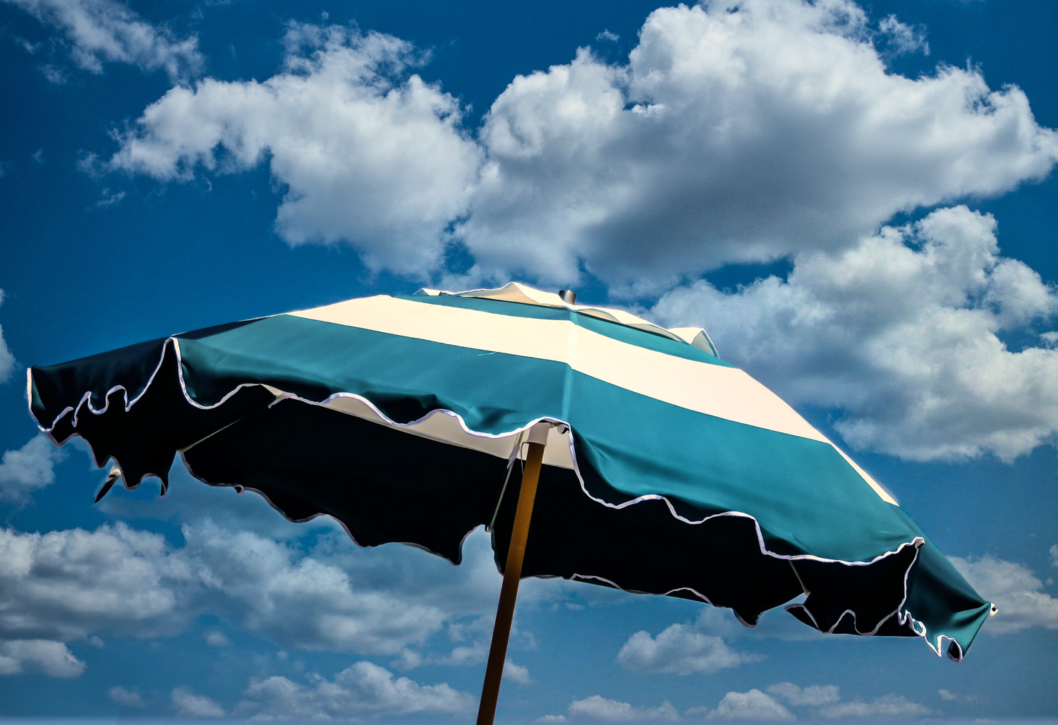 Teal and white striped beach umbrella against a bright blue sky with fluffy white clouds on a sunny day.