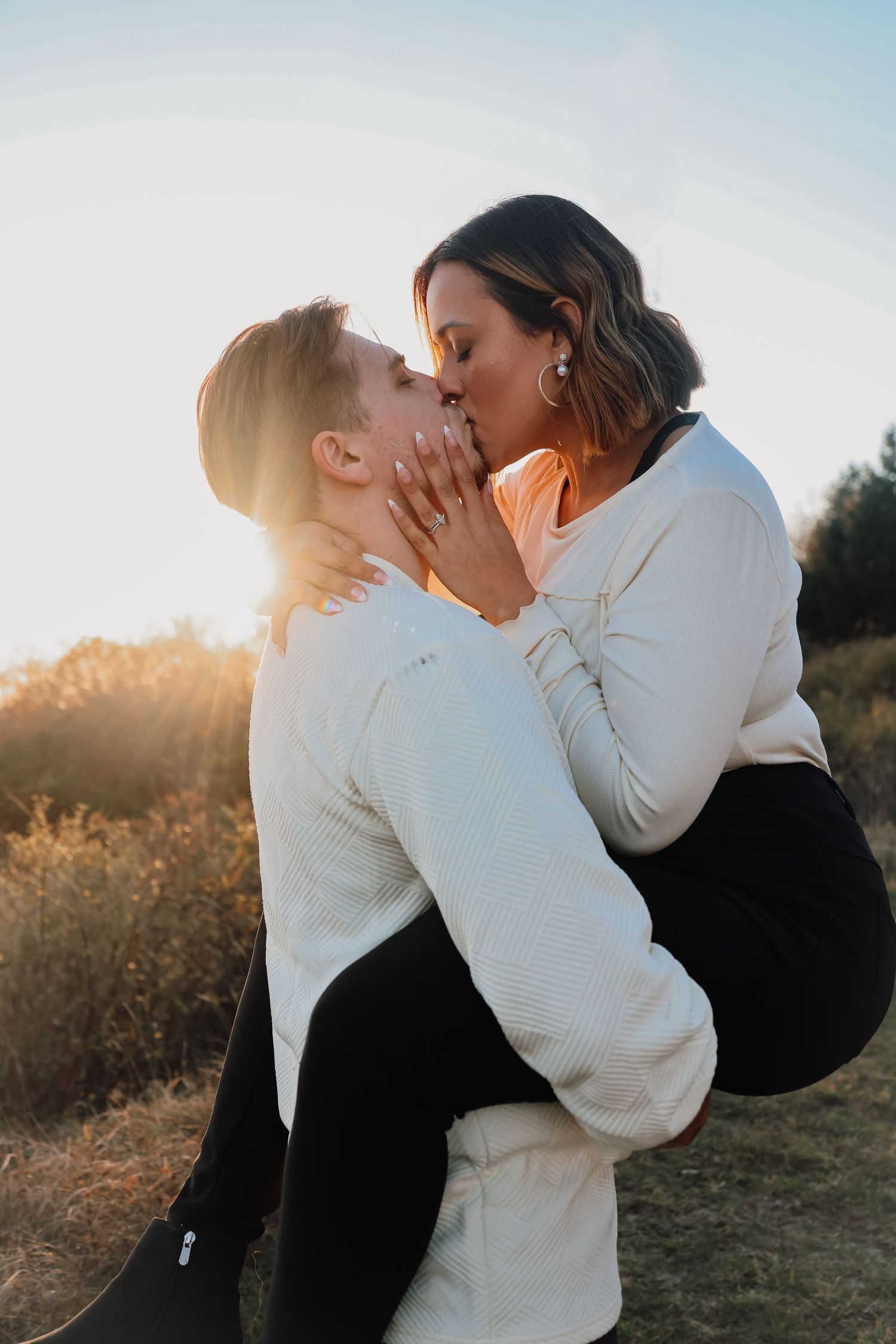 Couple sharing a romantic kiss at golden hour, man lifting woman in a field with warm sunset light behind them.