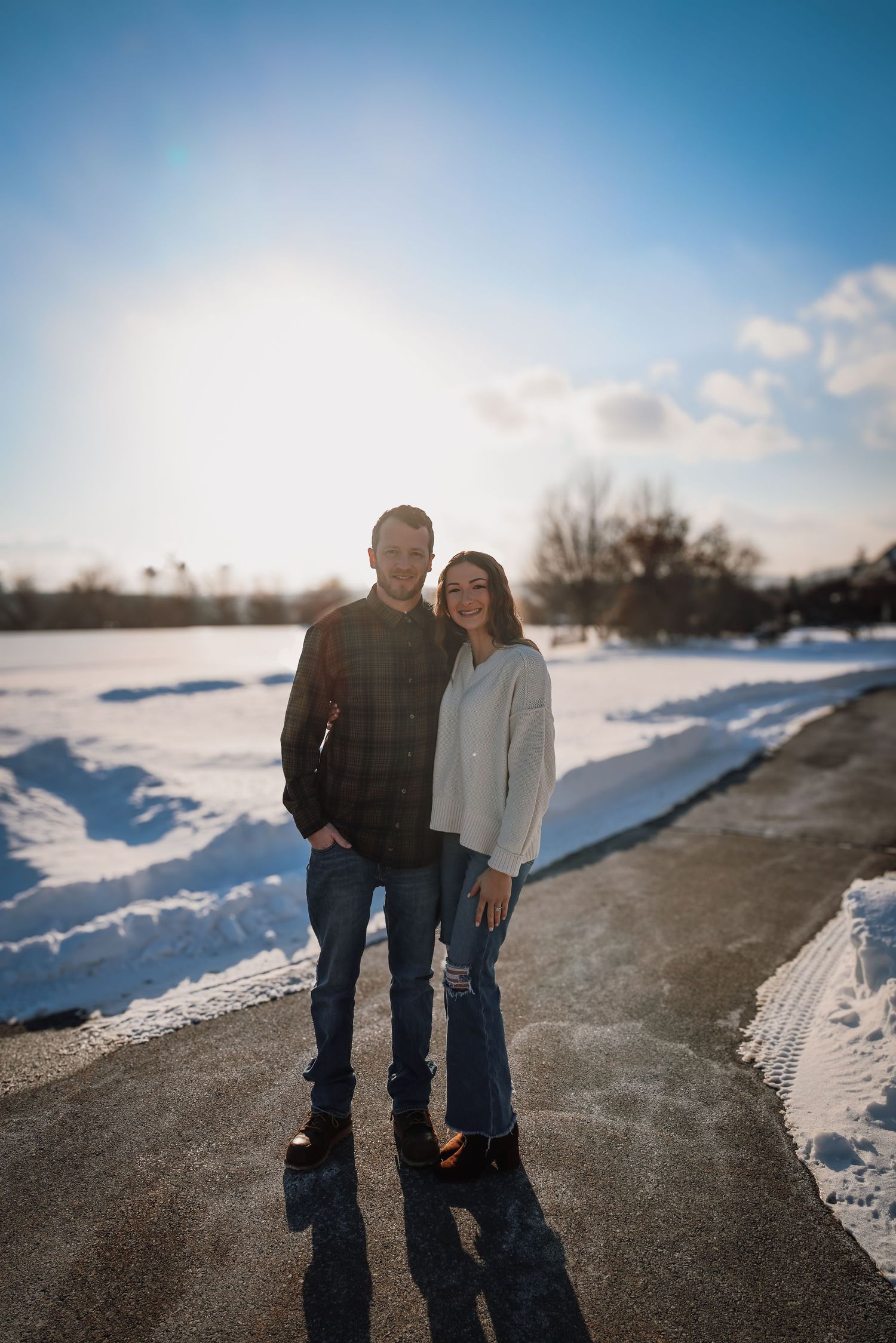 A couple stands together on a path during winter with snow-covered ground and blue sky in the background.