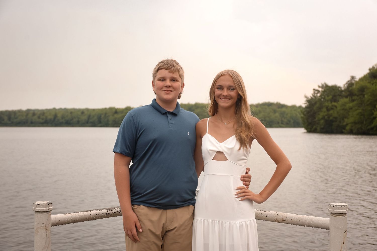 A couple poses together on a dock by a serene lake during sunset while holding wine glasses.