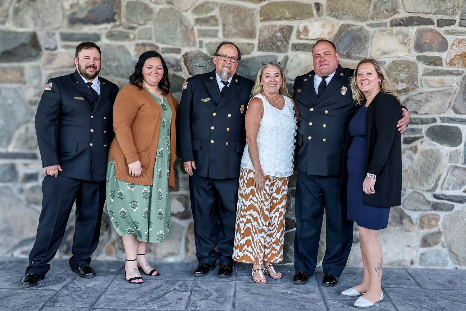 A group of people in formal attire and military uniforms pose together against a stone wall background.
