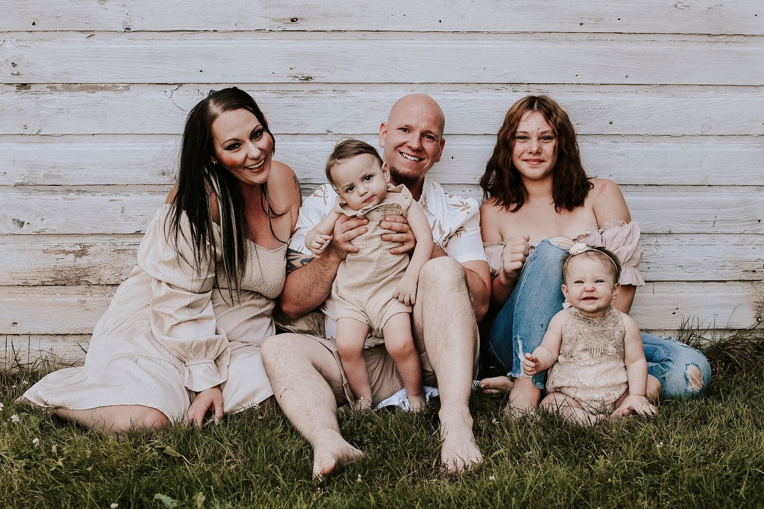Family sitting casually in a row against weathered white wooden wall backdrop.