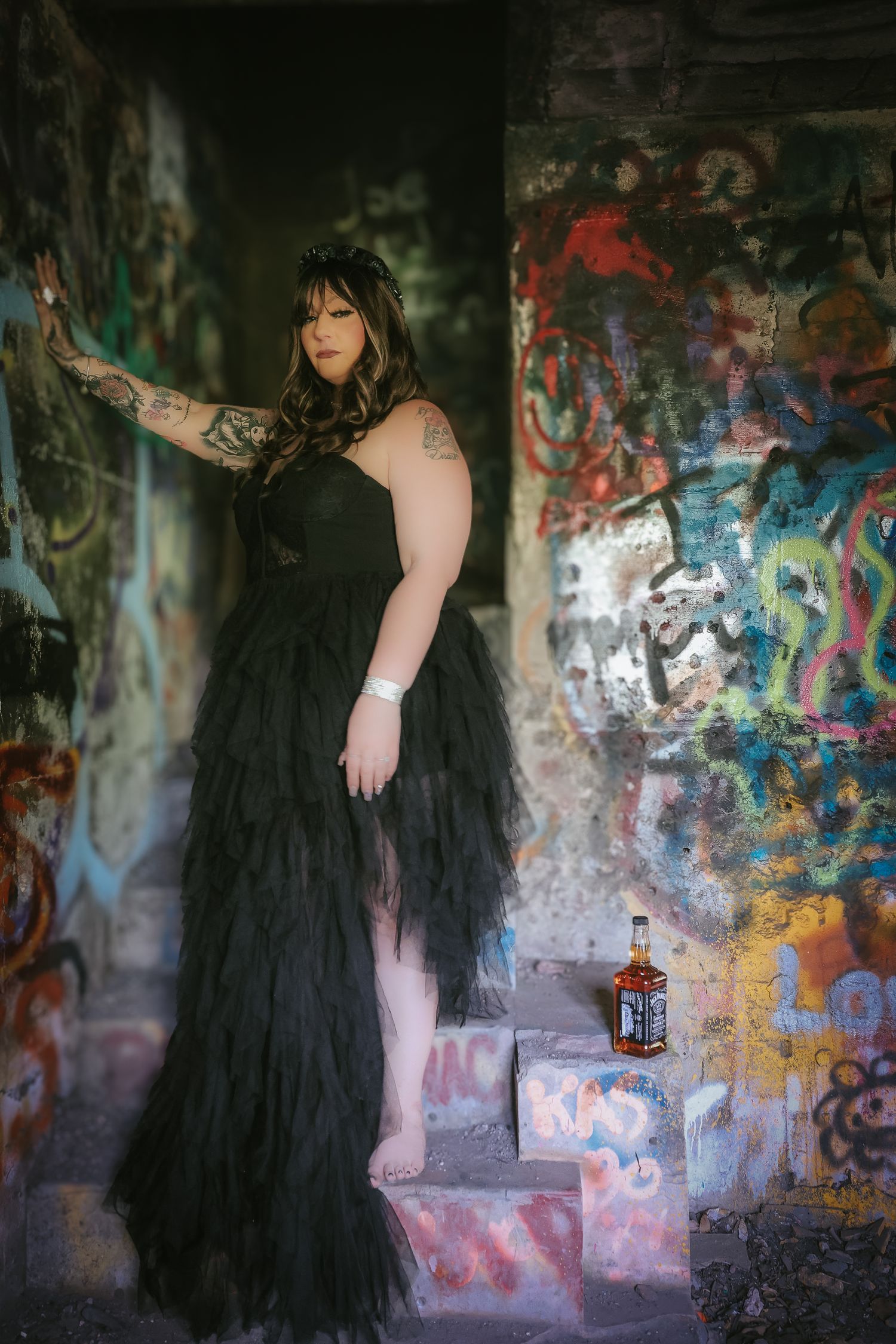 A figure in a dramatic black tulle gown poses against a graffiti-covered wall in an abandoned urban setting.