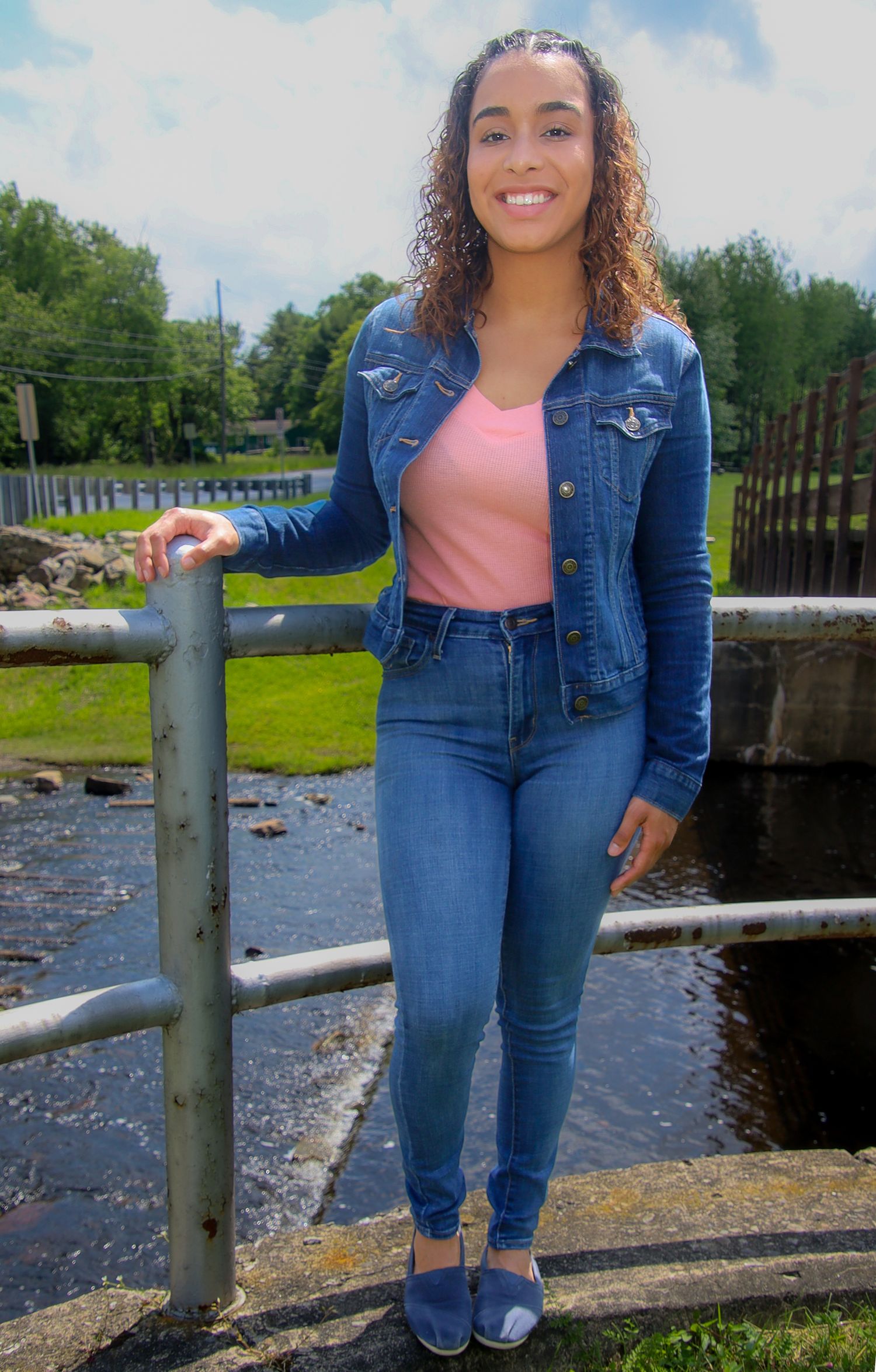 Woman in denim jacket and jeans poses near waterfront railing on sunny day.