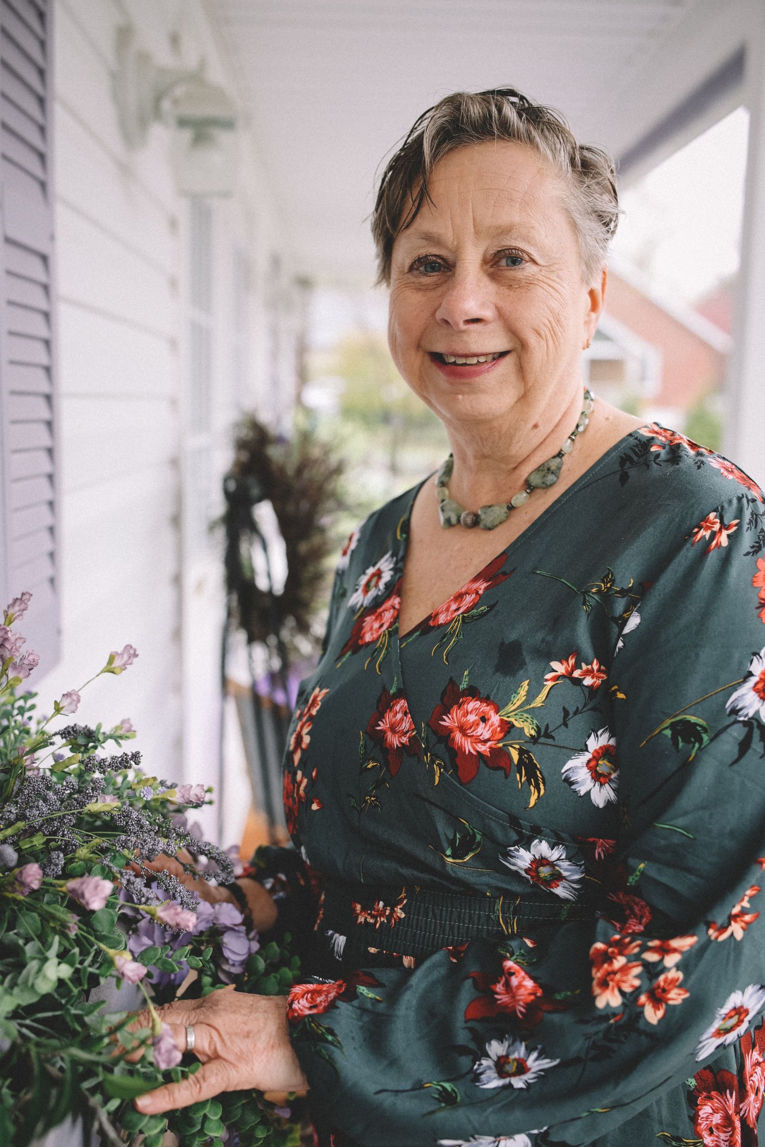 Woman in green floral dress stands on porch surrounded by potted plants and flowers.
