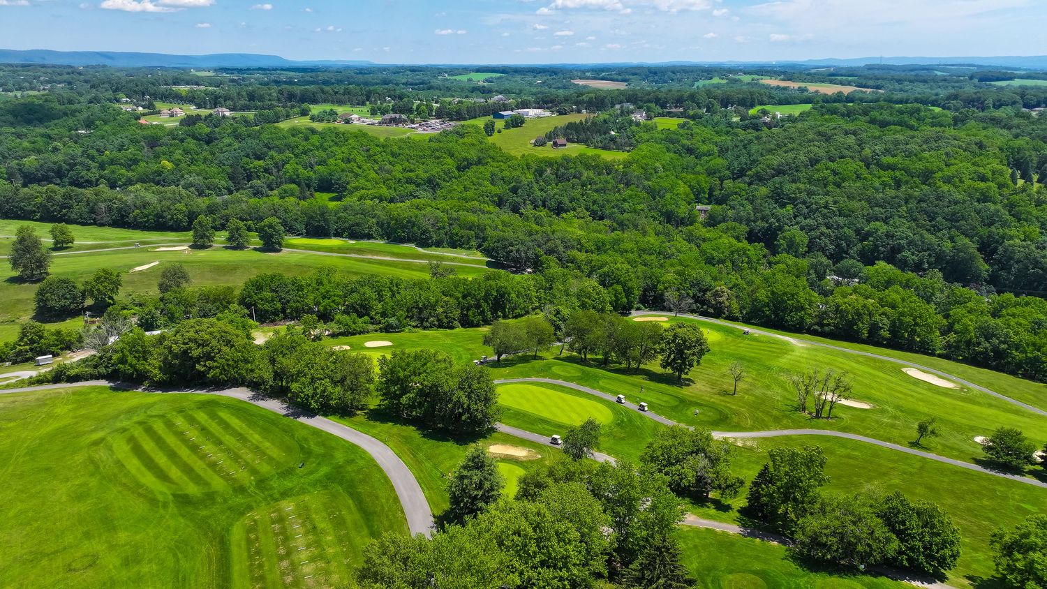 Aerial panoramic view of lush green golf course with rolling fairways and tree-lined borders on a sunny day.