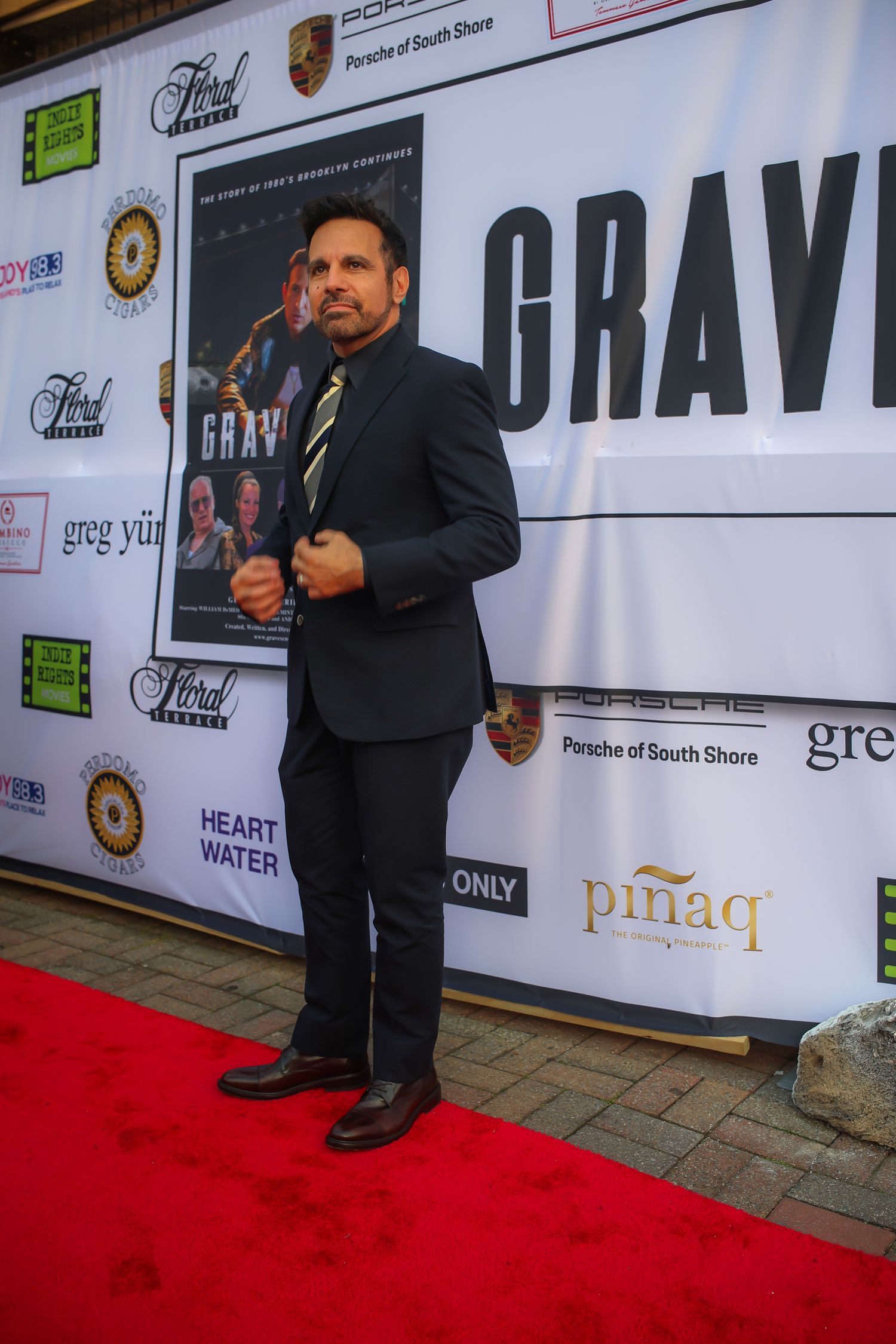 A person wearing a dark suit stands on a red carpet in front of a backdrop with sponsor logos at an event.
