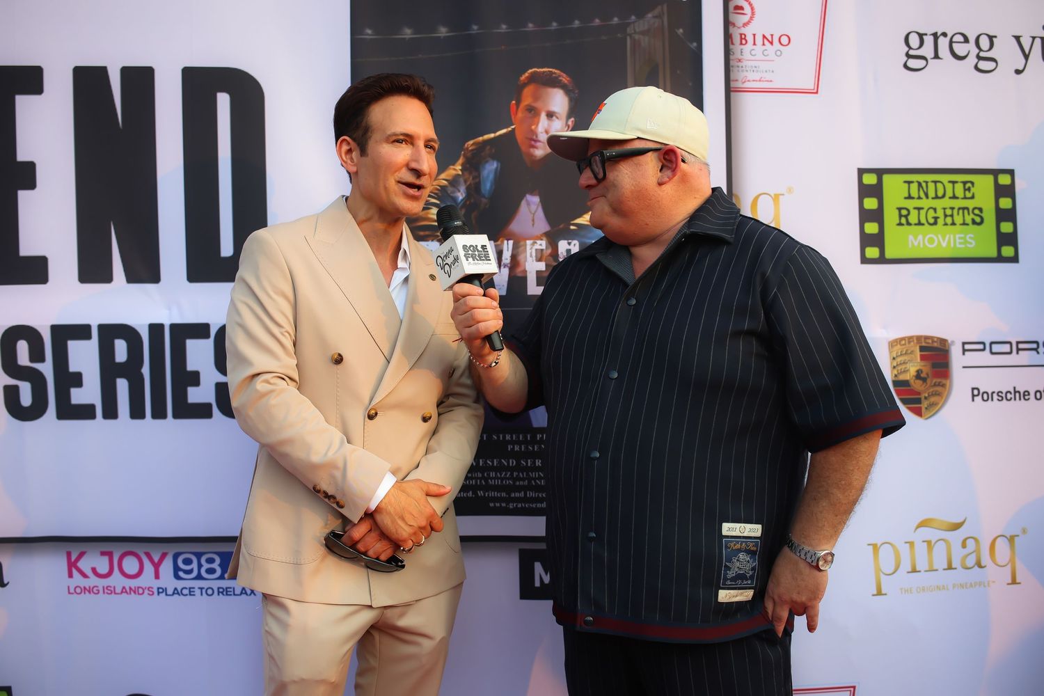Two people stand together at a red carpet event with a backdrop showing sponsor logos and text.