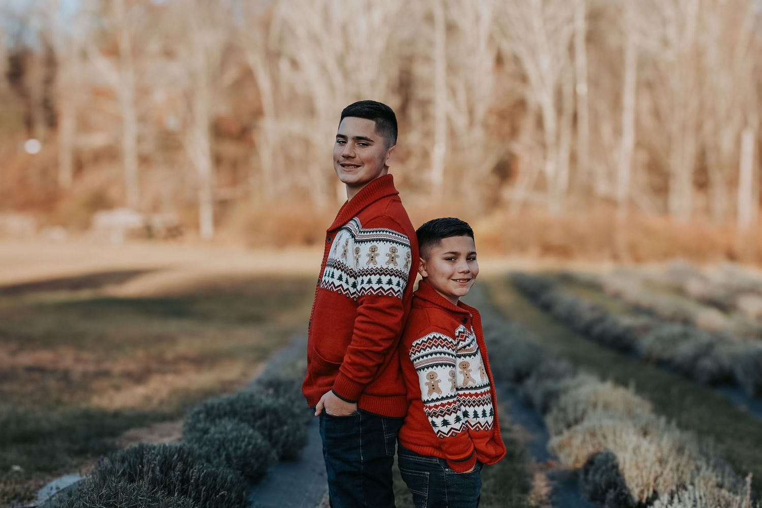 Two people wearing matching red Christmas sweaters stand together in a wooded outdoor setting during winter.