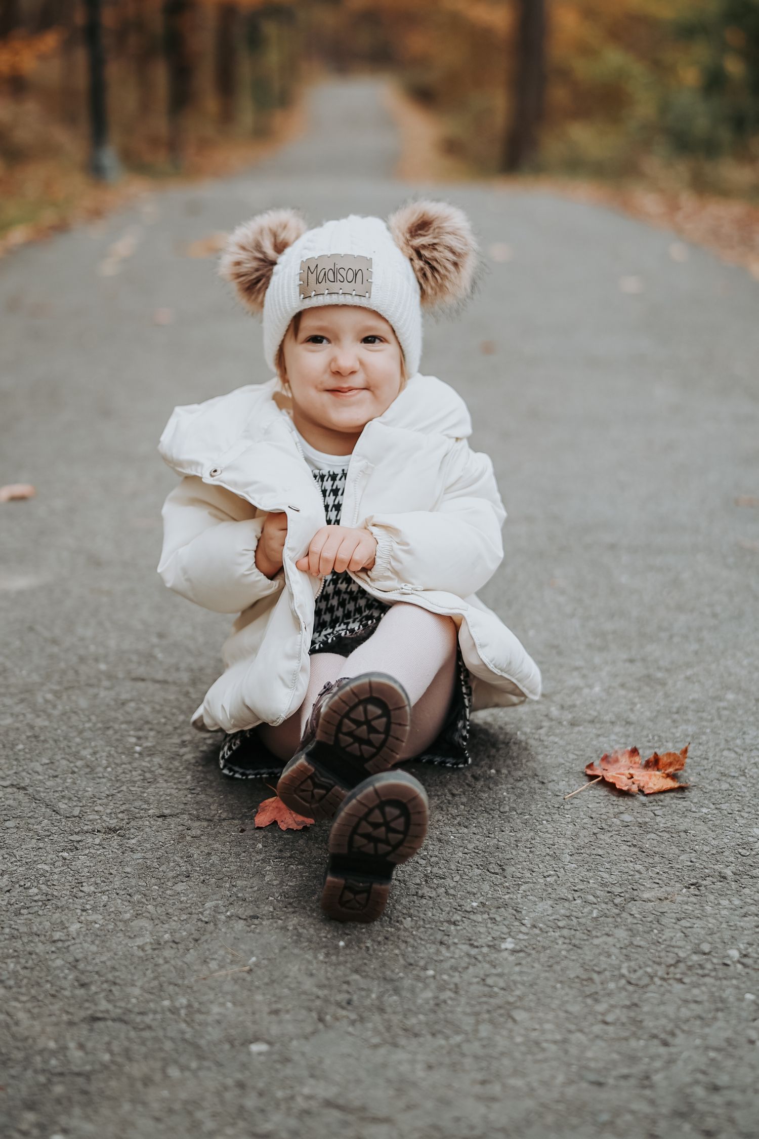 A small child in a white dress and winter hat lies on an autumn path covered in fallen leaves.