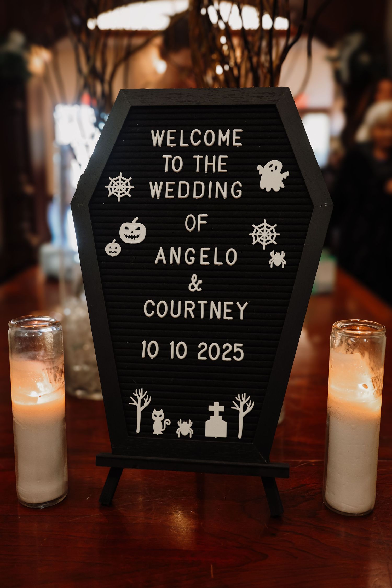 Black coffin-shaped welcome sign with spooky Halloween themed decorations for a wedding celebration, illuminated by white candles.