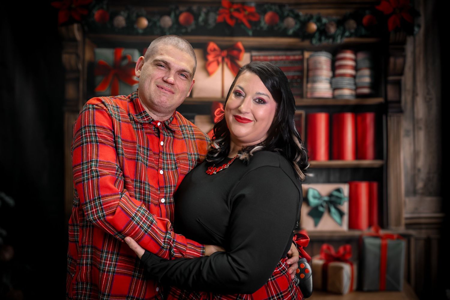 A couple in coordinating red plaid and black outfits pose together for holiday portraits in a festive setting.