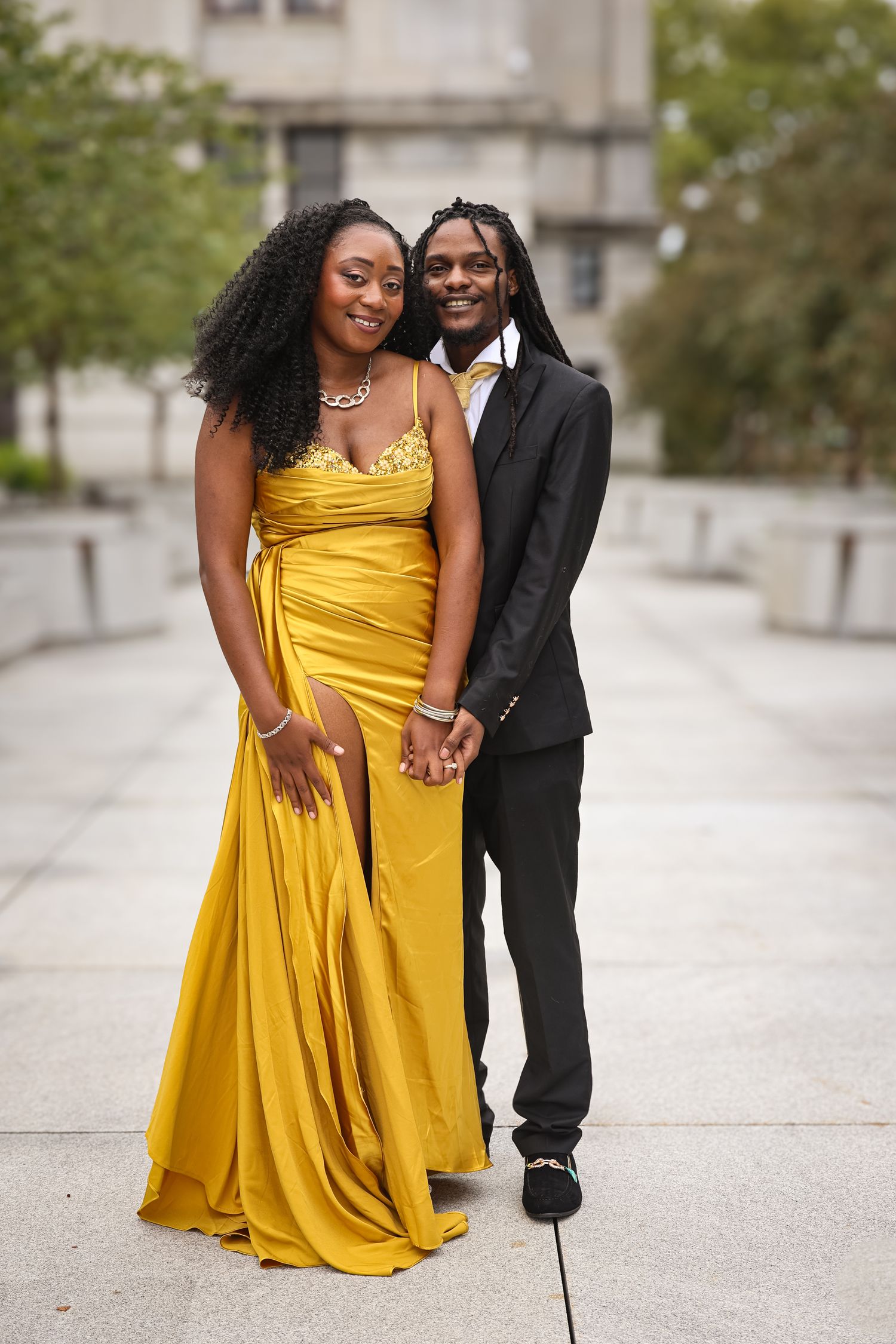 A couple poses together outdoors, with one wearing a flowing yellow gown and the other in a black suit.