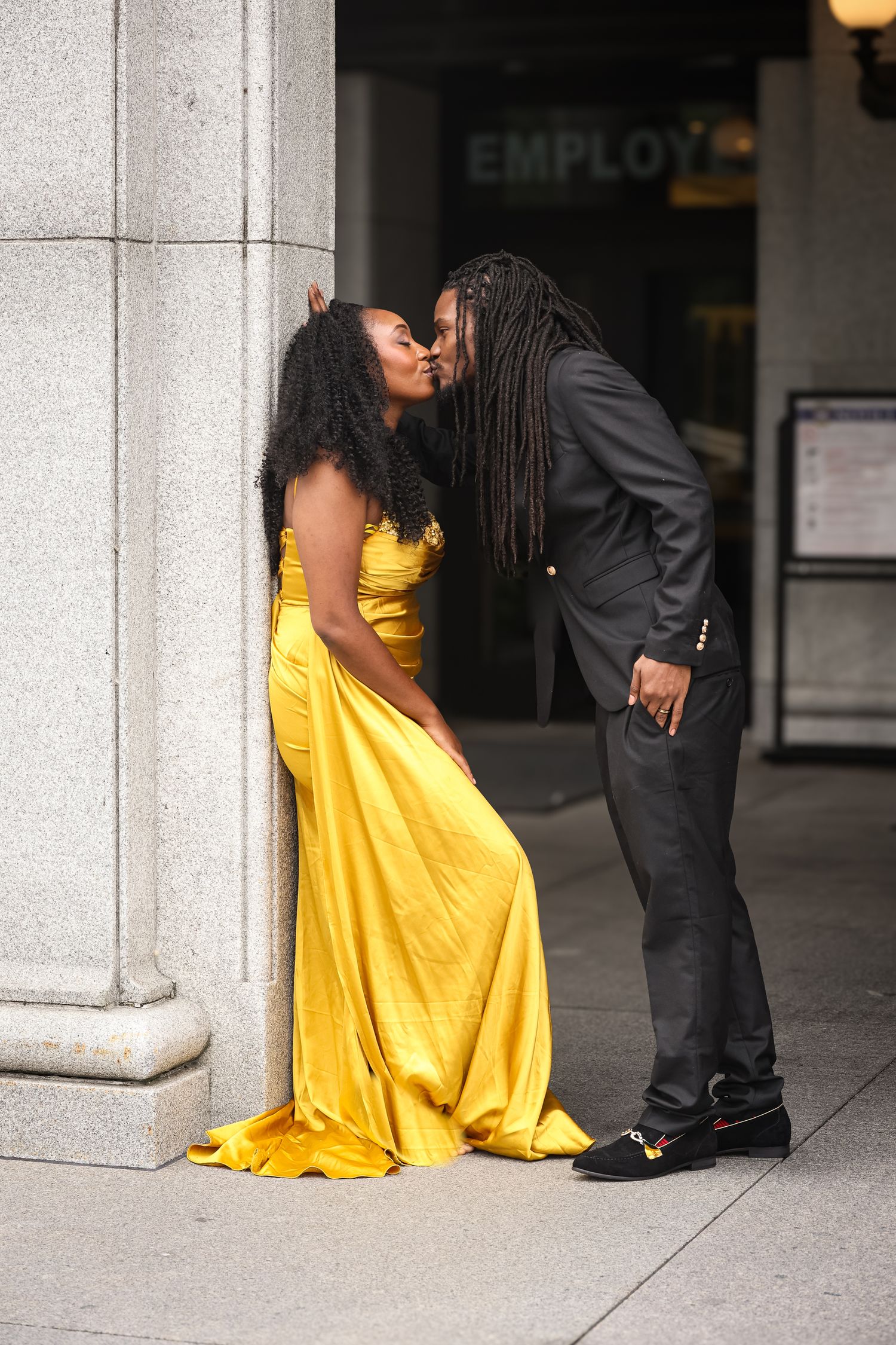 A couple in formal attire shares a tender moment against a stone column, with one wearing a flowing yellow dress.