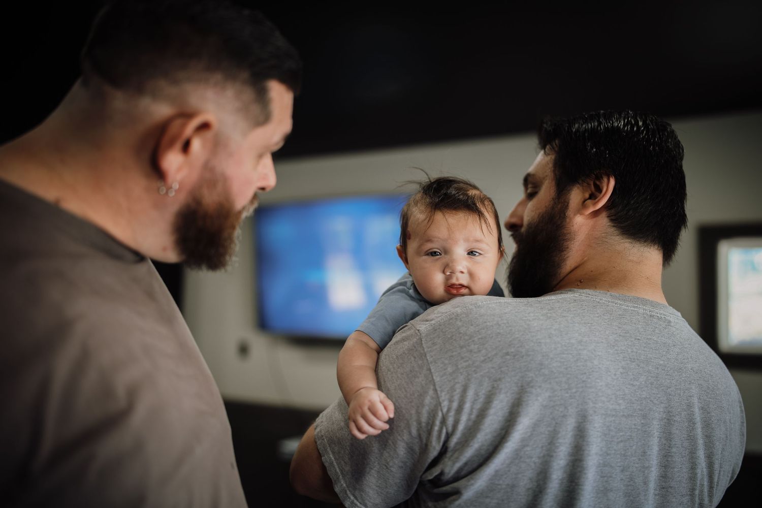 Two adults in gray shirts hold a small baby while having a conversation in an indoor setting.