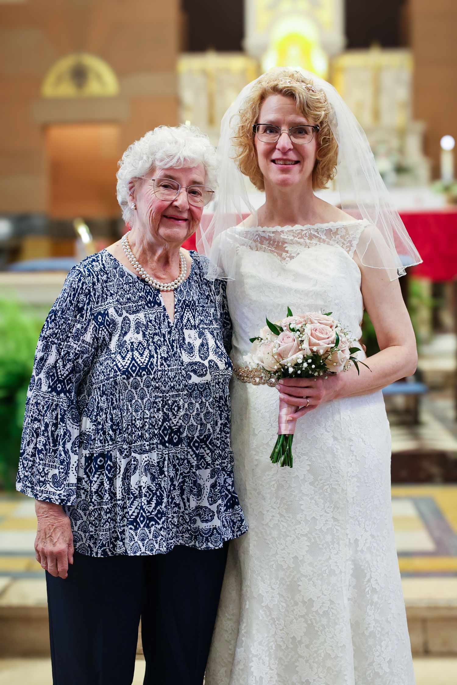 A bride in a white lace dress poses with an elderly woman wearing a navy and white patterned blouse at a wedding.