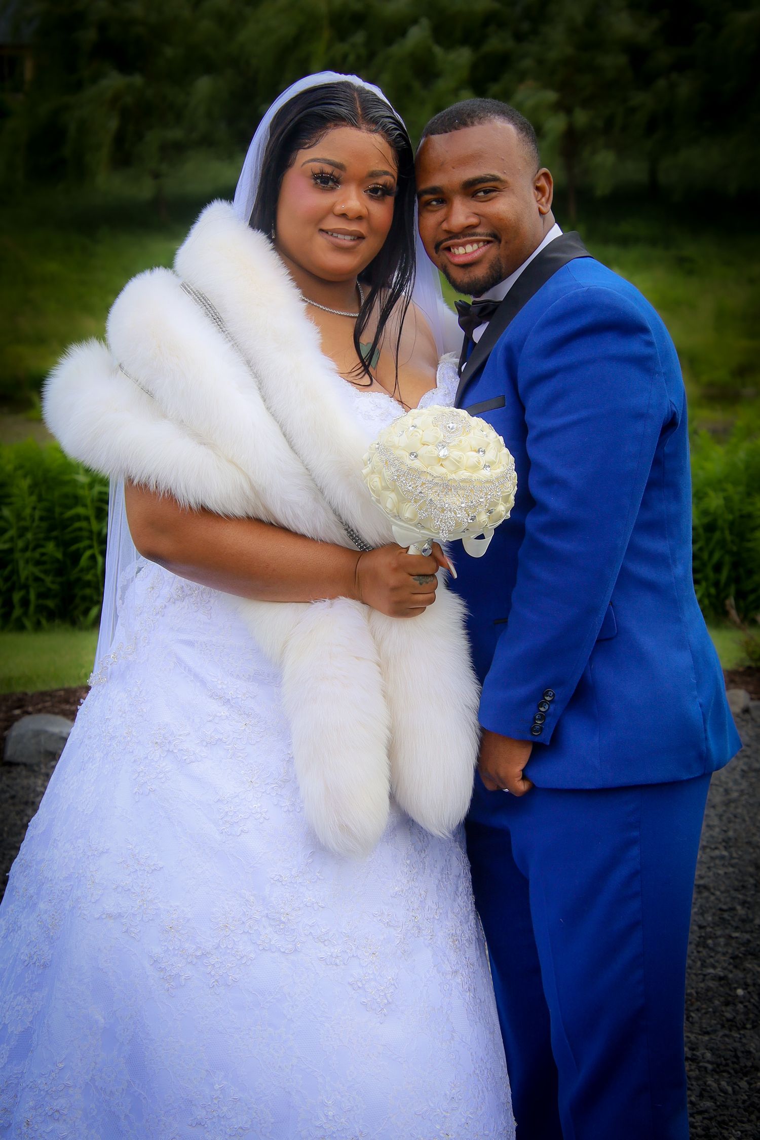 A couple in wedding attire poses outdoors, with the bride in a white gown and fur stole and groom in royal blue suit.